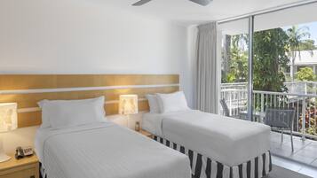 A hotel room with two beds, a ceiling fan, a balcony with a view of greenery, and a nightstand with a lamp and phone.