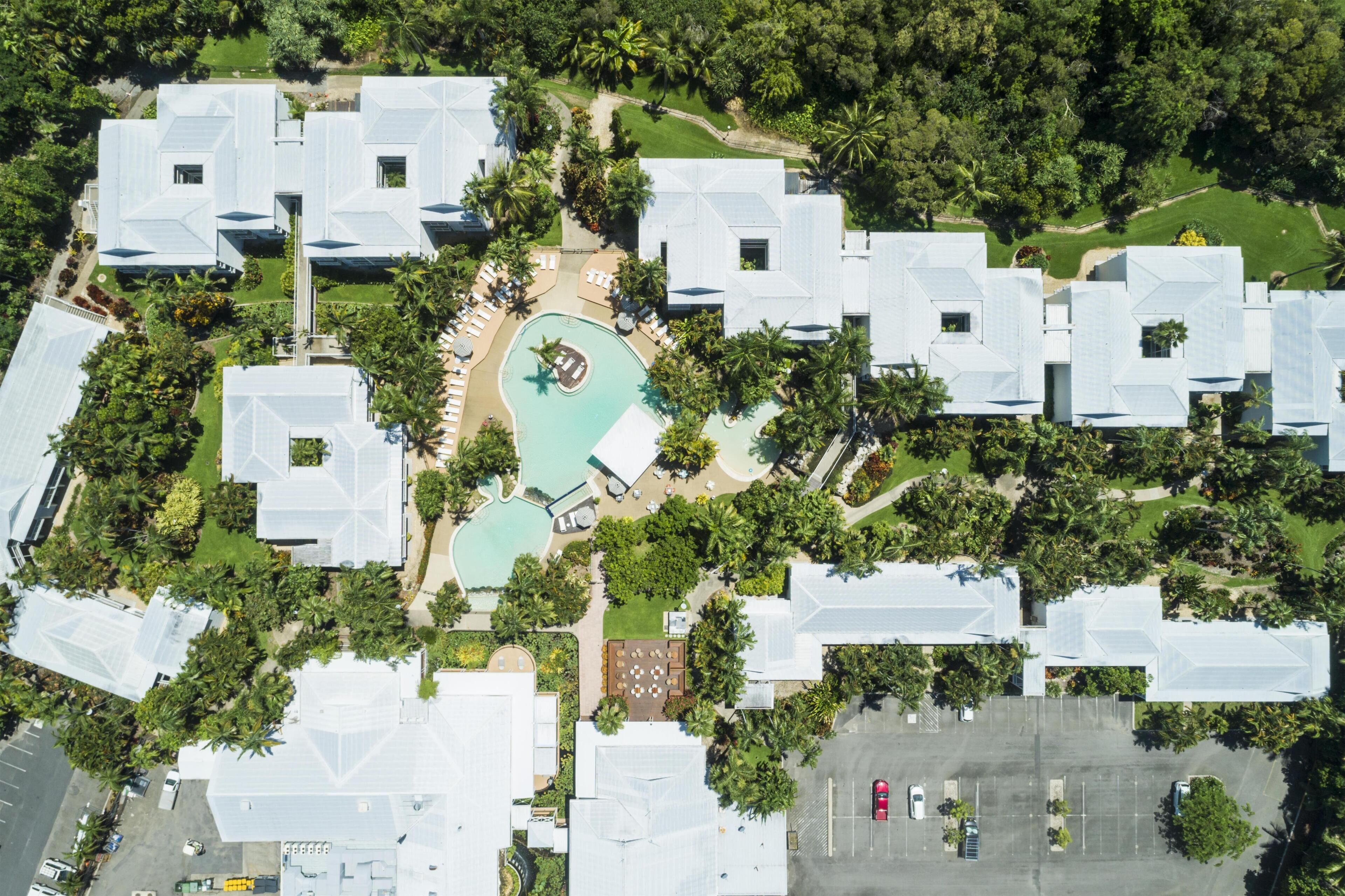 An aerial view of a resort with multiple white-roofed buildings, a central swimming pool, and surrounding greenery.
