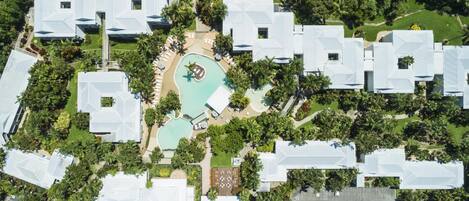 An aerial view of a resort with multiple white-roofed buildings, a central swimming pool, and surrounding greenery.