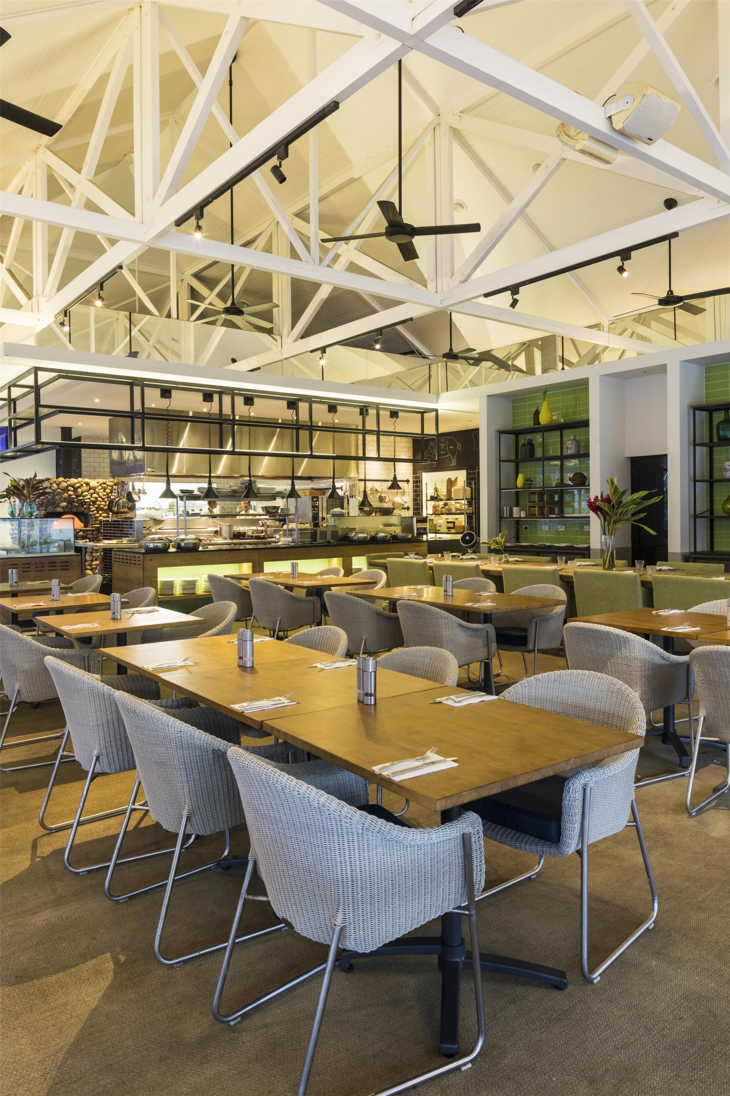 A spacious dining area with high ceilings and modern lighting fixtures. Tables are arranged for groups, and the space features a mix of seating options.