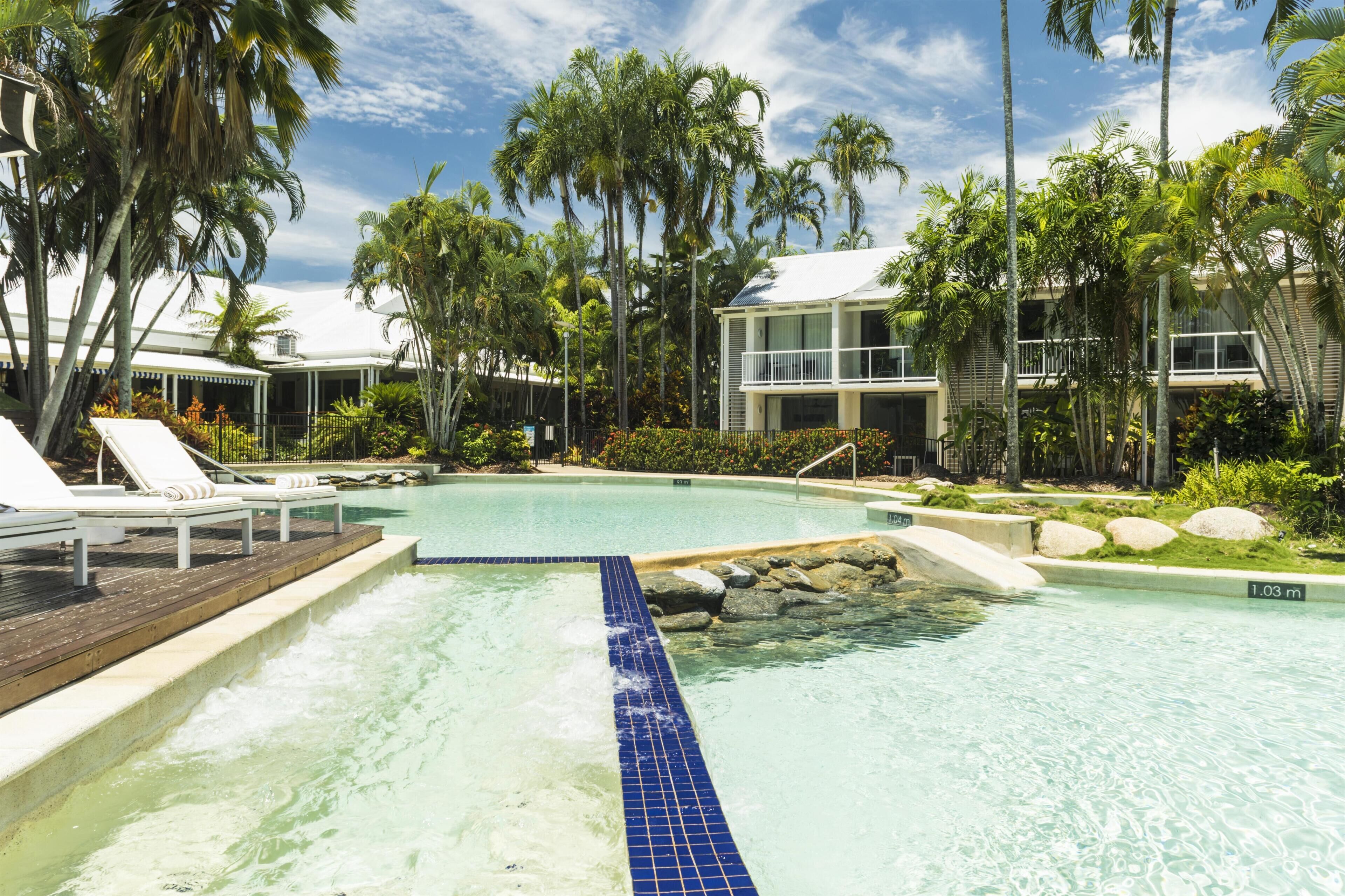 Una piscina con sillones de descanso, rodeada de palmeras y un edificio con balcones.