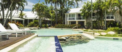 Una piscina con sillones de descanso, rodeada de palmeras y un edificio con balcones.