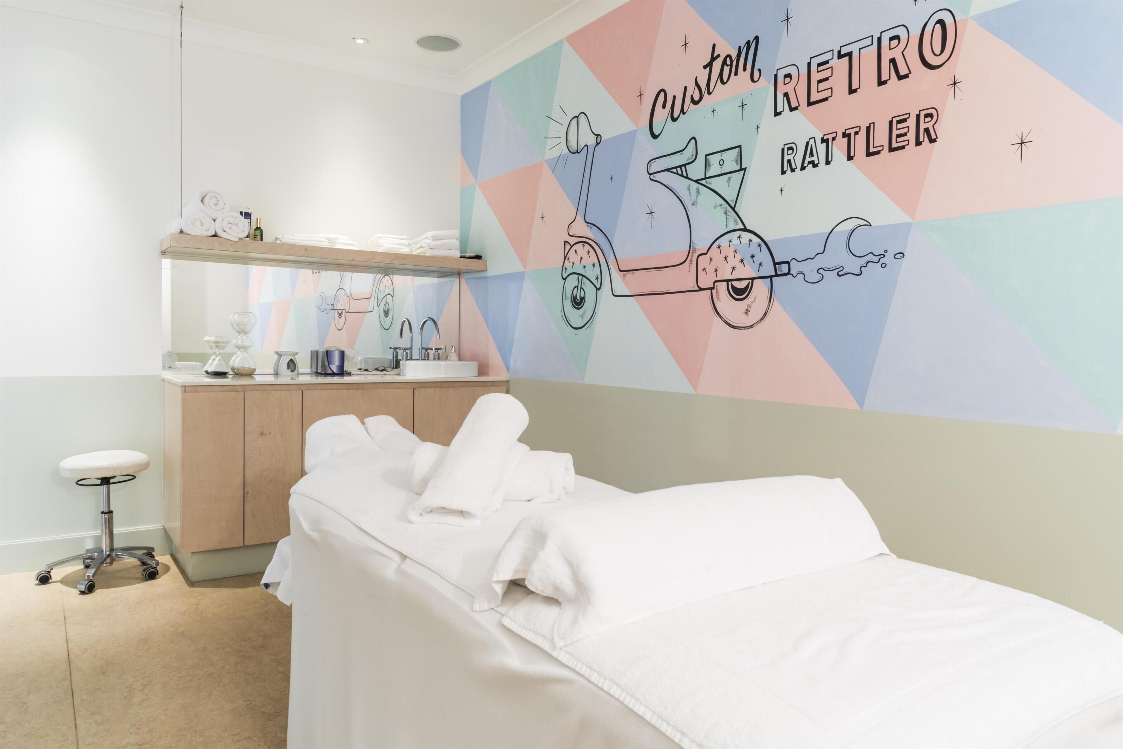 Een hotelkamer met een bed, een bureau met stoel, een spiegel en een wand met een kleurrijk geometrisch ontwerp.