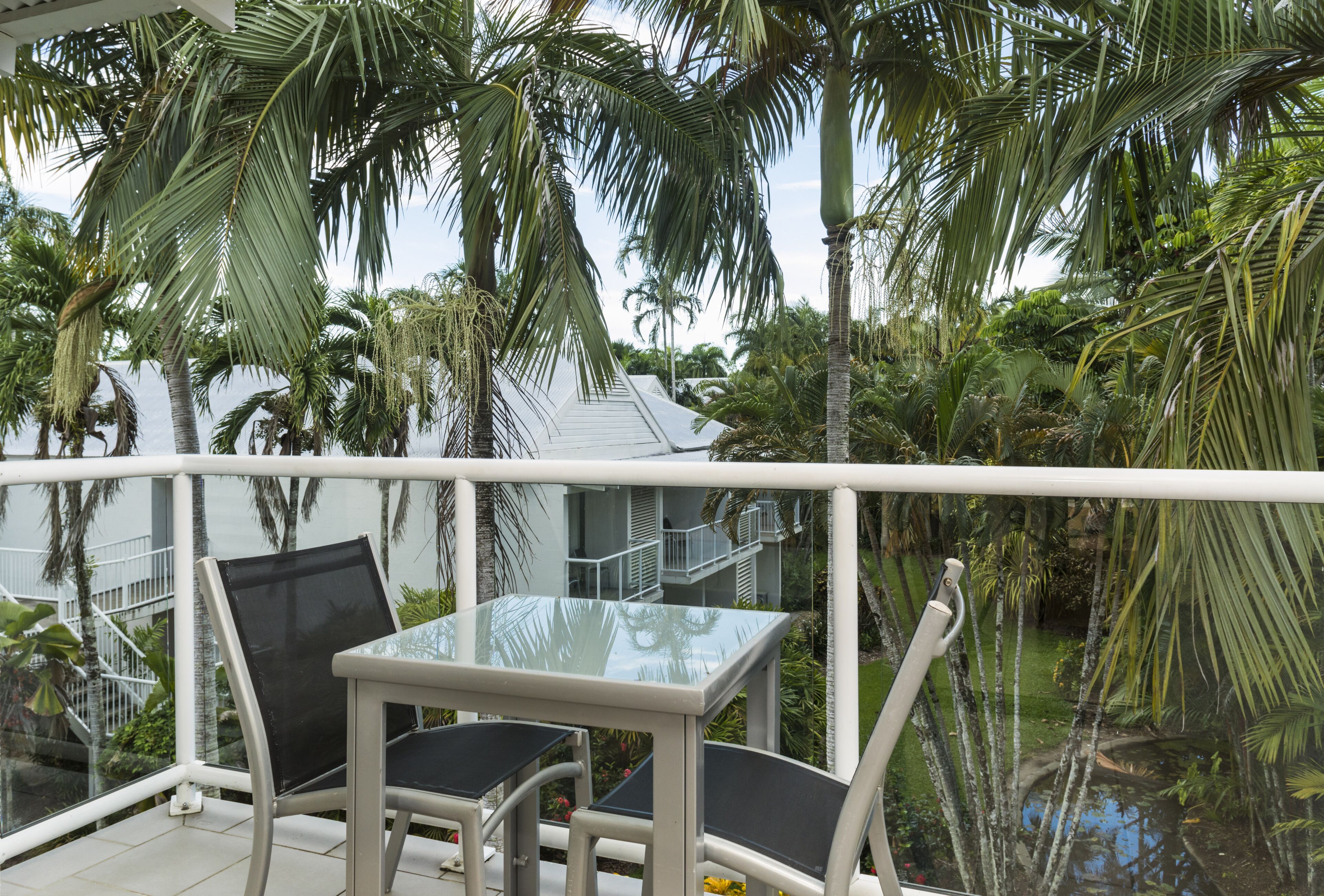 A balcony with a glass table and chairs, surrounded by palm trees and a view of a building with balconies.