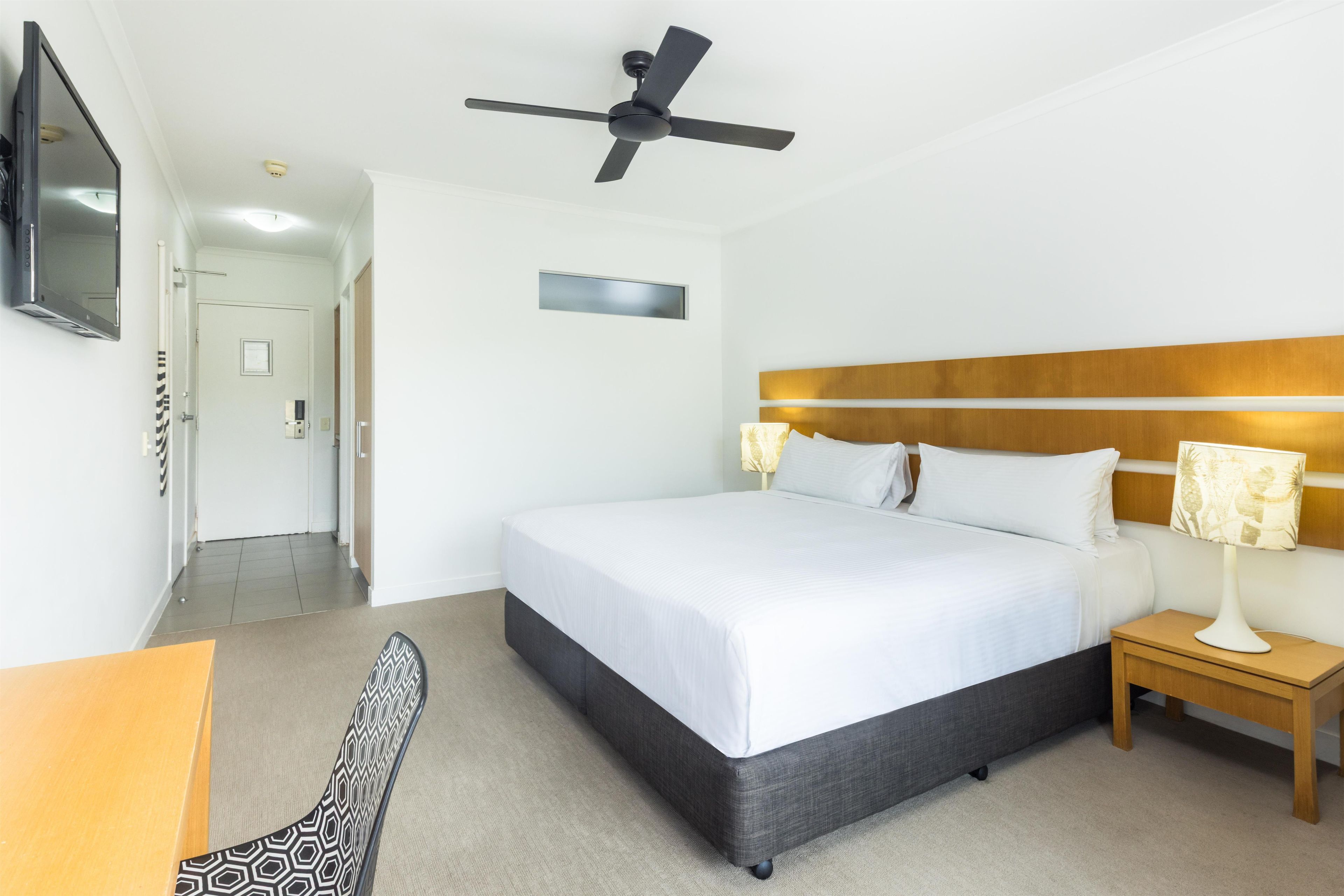 Habitación de hotel con cama, mesita de noche, silla, ventilador de techo, televisor y una puerta que da a otra habitación.