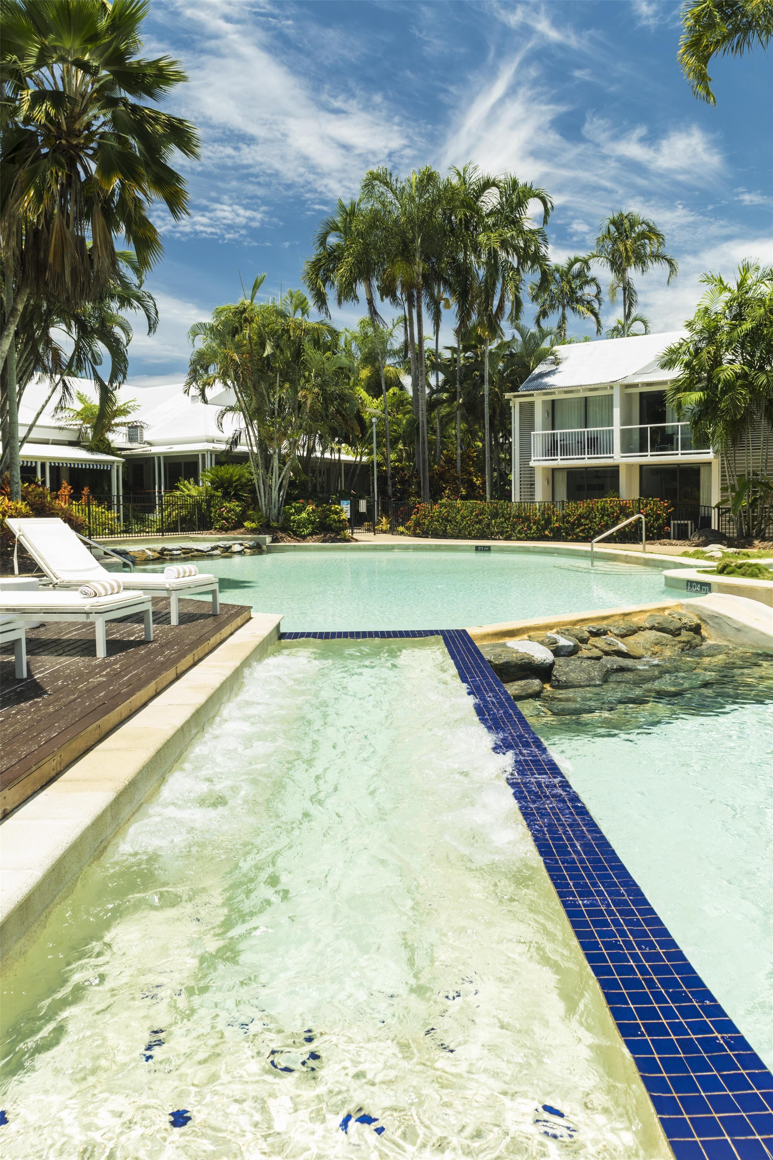 Una piscina con sillones, palmeras y un edificio con balcón.