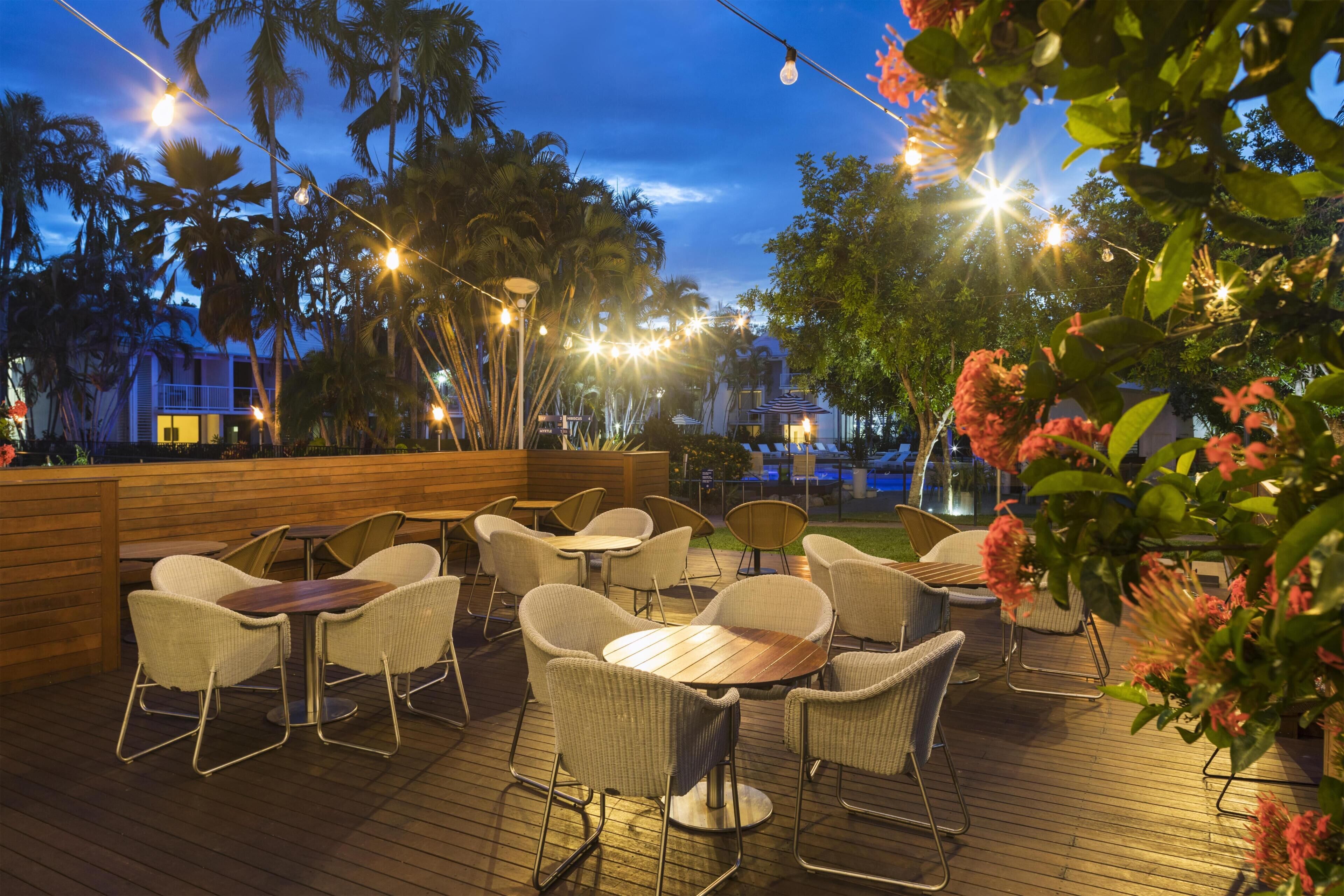 An outdoor patio with wicker chairs and tables, string lights, and palm trees.