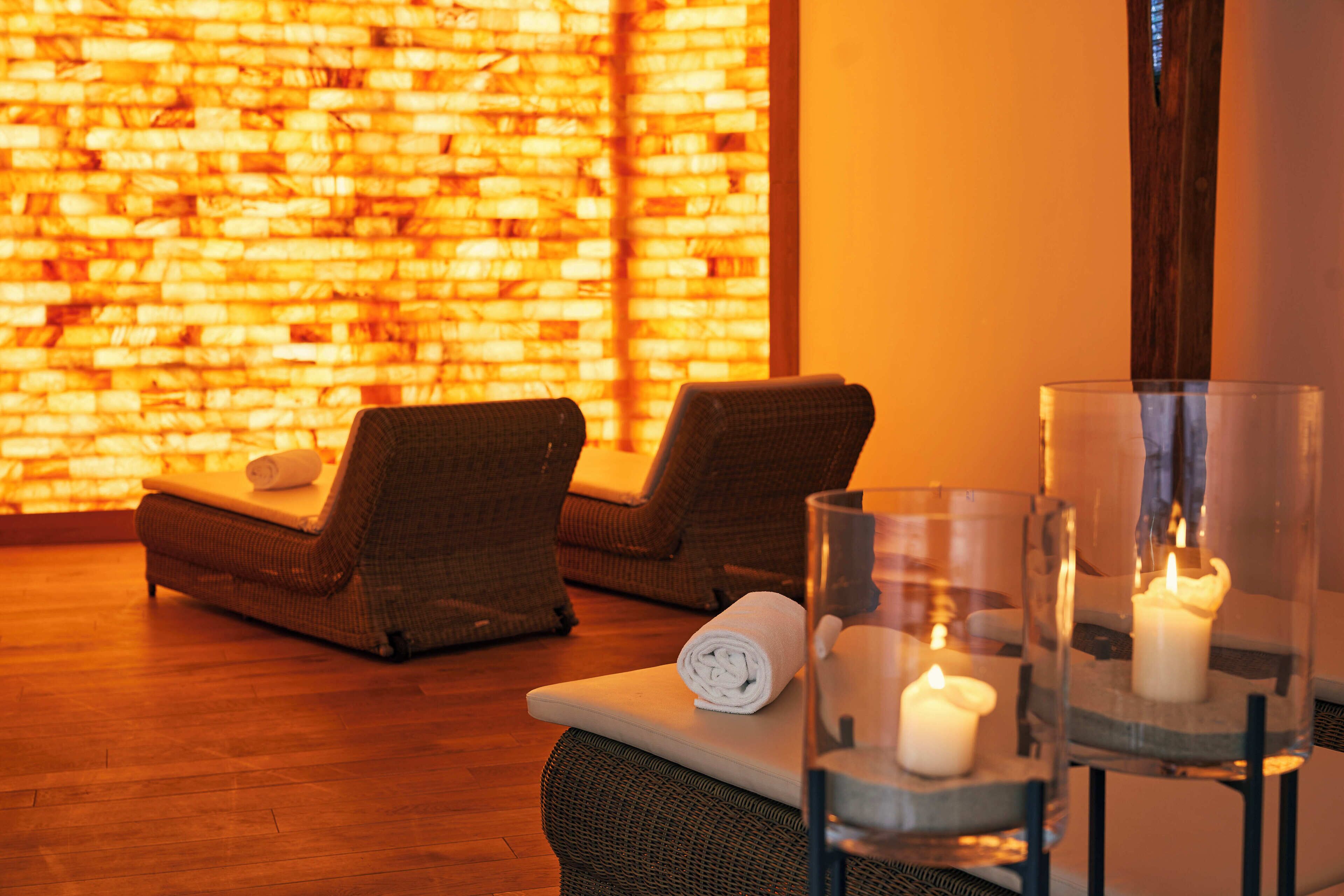 Um espaço de spa com duas poltronas de vime, uma mesa de vidro com toalha enrolada e velas em recipientes de vidro.