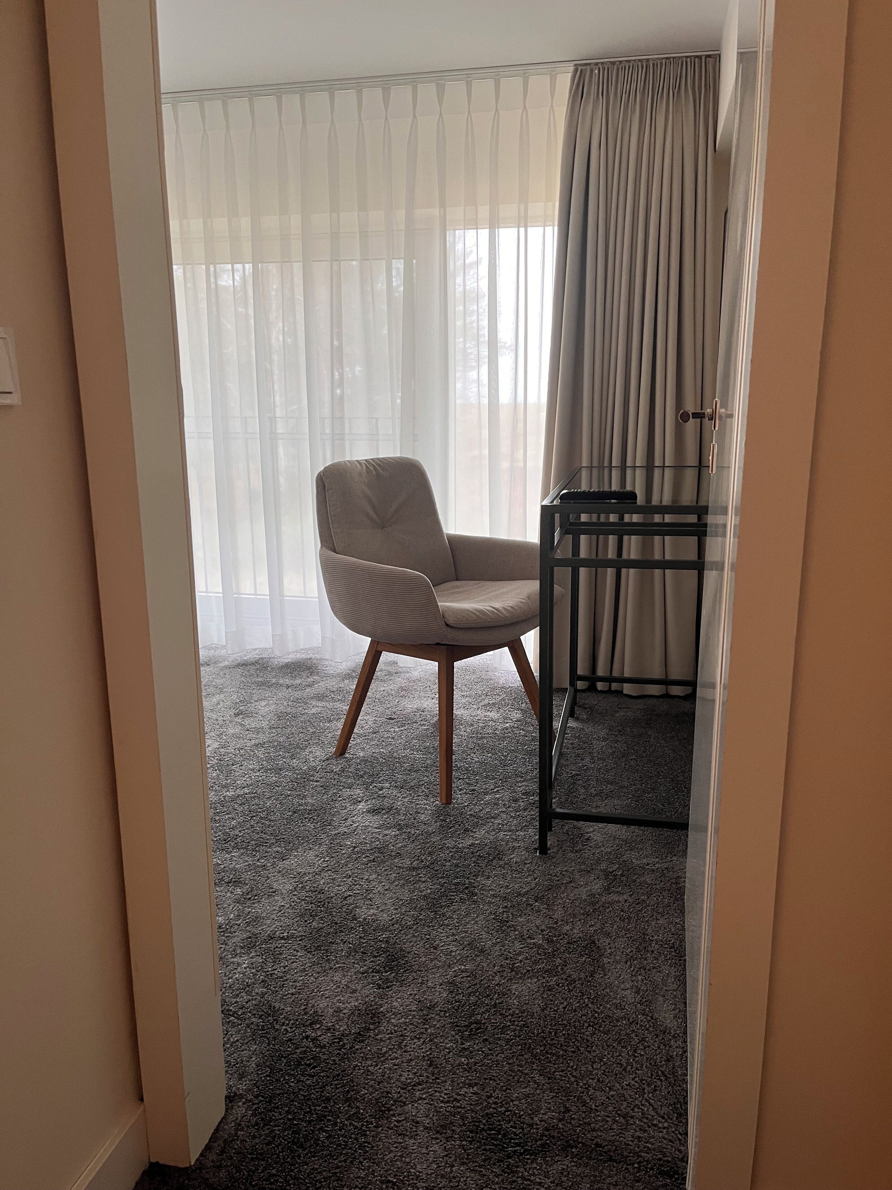 A room with a grey carpet, a beige armchair, a small table, and a window with sheer curtains.