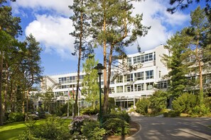 Un bâtiment moderne à la façade vitrée, entouré d’un jardin bien entretenu où l’on trouve différentes espèces d’arbres et d’arbustes.