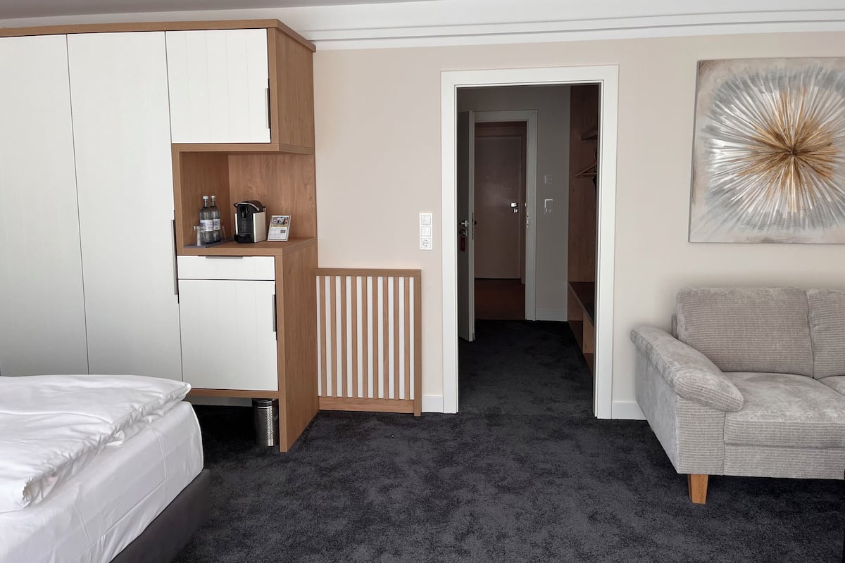 Ein Hotelzimmer mit einem Bett, einem Kleiderschrank, einer Küchenzeile, einer Heizung, einer Tür zu einem anderen Zimmer und einem grauen Sofa.