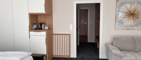 A hotel room with a bed, a wardrobe, a kitchenette, a radiator, a door to another room, and a grey sofa.