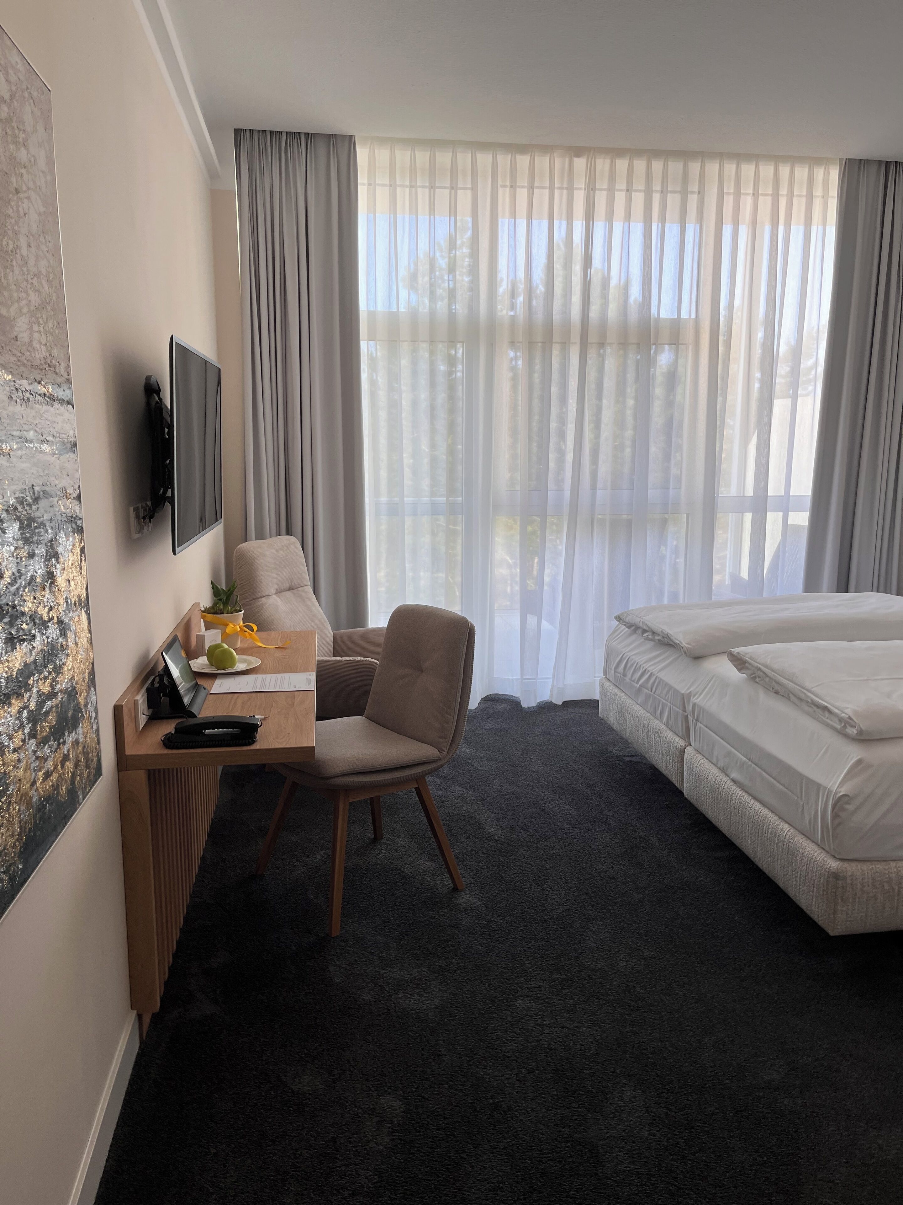 A hotel room with a bed, a desk with a chair, a flat-screen TV, and a large window with sheer curtains.