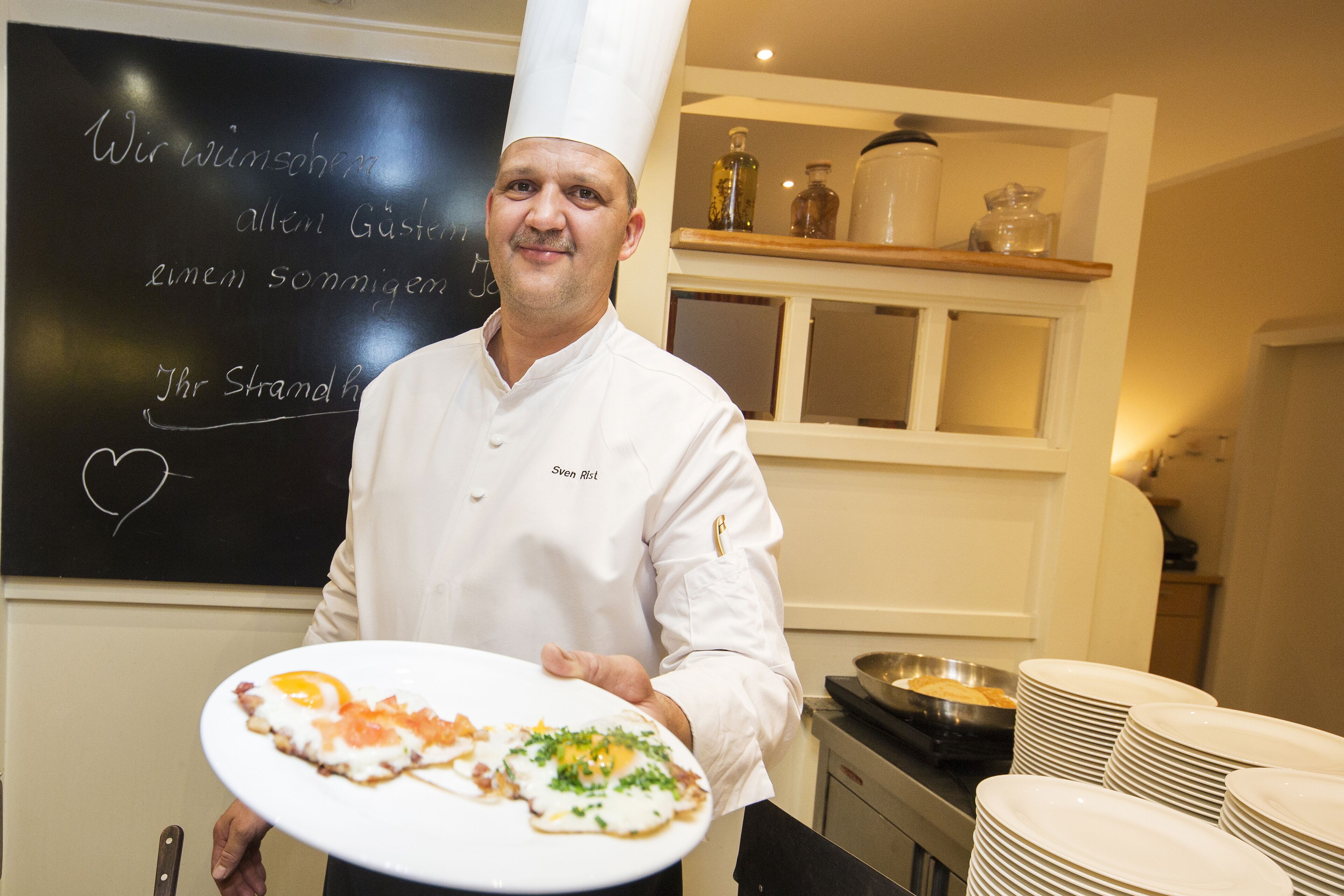 A chef in a white uniform holding a plate with food, standing in a kitchen with a chalkboard in the background.