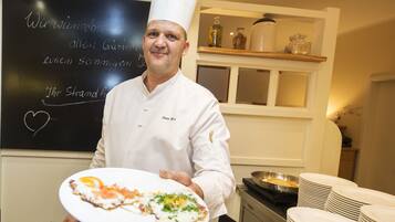 A chef in a white uniform holding a plate with food, standing in a kitchen with a chalkboard in the background.