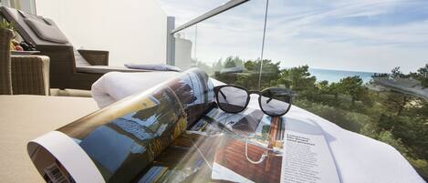 A magazine on a table with a pair of glasses, a wicker chair, and a view of the sea.