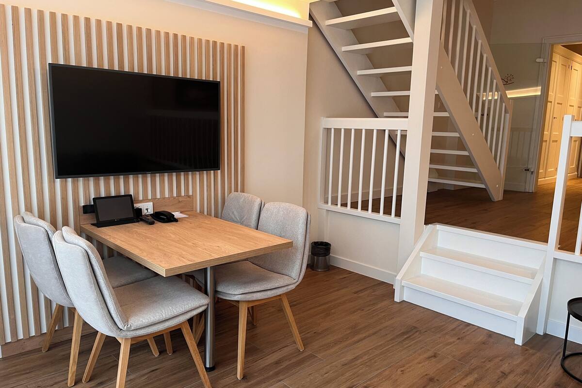 Ein modernes Wohnzimmer mit einem Holzesstisch, grau gepolsterten Stühlen, einem Flachbildfernseher an einer Wand aus Holzpaneelen und einer Treppe, die zu einer oberen Ebene führt.