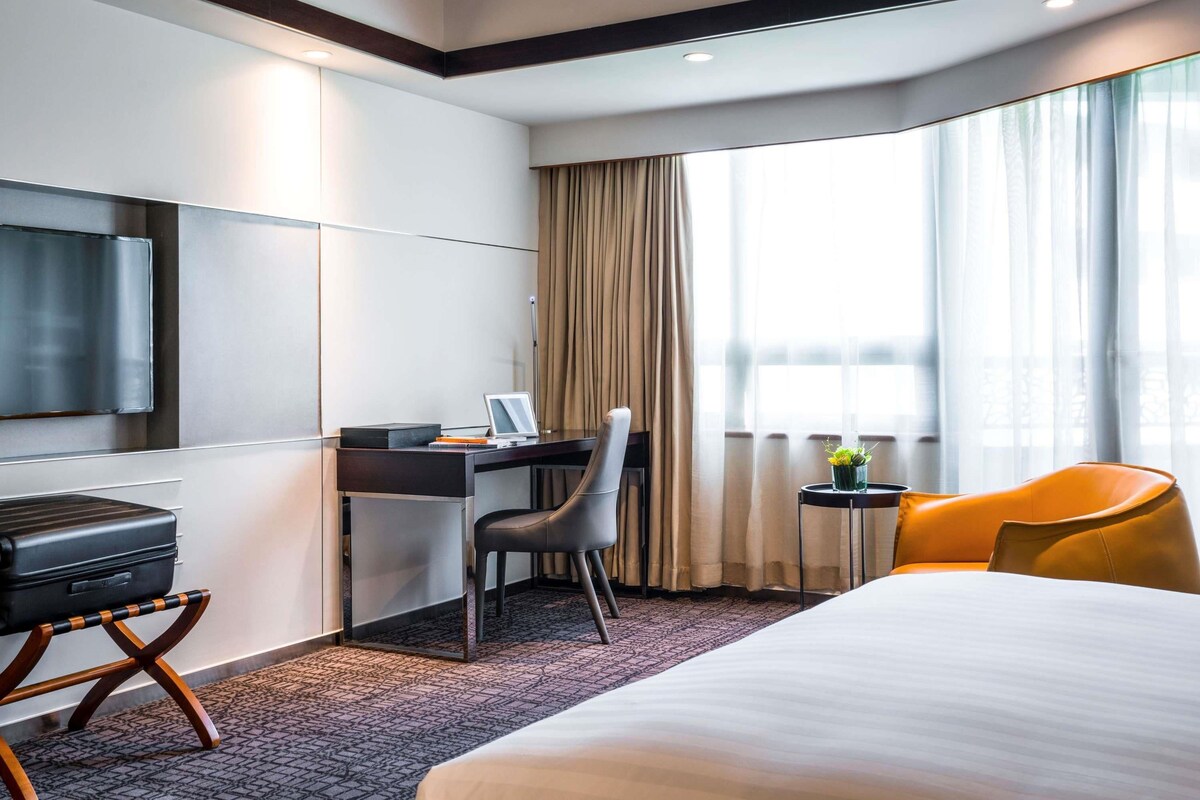 Ein Hotelzimmer mit Bett, Schreibtisch, Stuhl, Fernseher und einem Fenster mit Vorhängen.