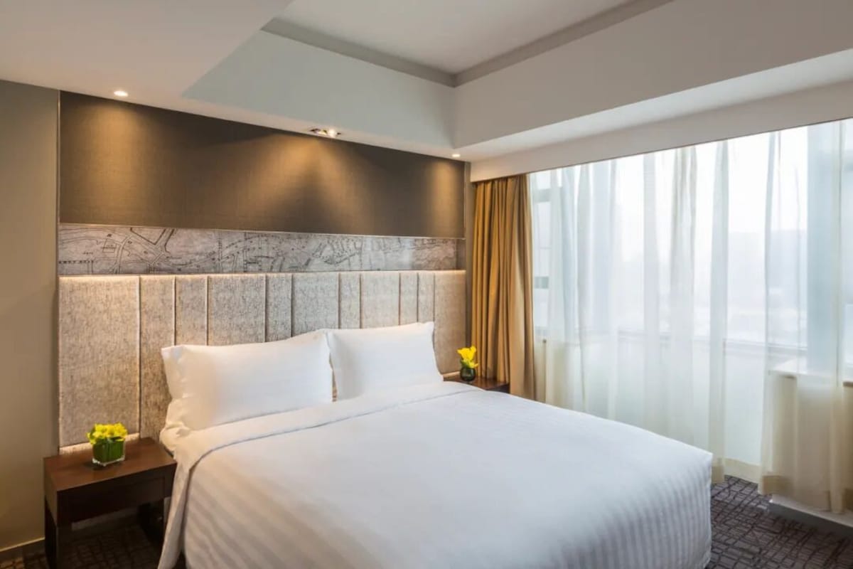 Ein Hotelzimmer mit einem großen Bett, einem Marmorkopfstück, einem Nachttisch mit Blumenvase und einem großen Fenster mit Vorhängen.