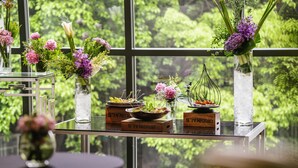 Une table ornée de vases de fleurs et d’un saladier, placée devant une fenêtre donnant sur un paysage verdoyant.