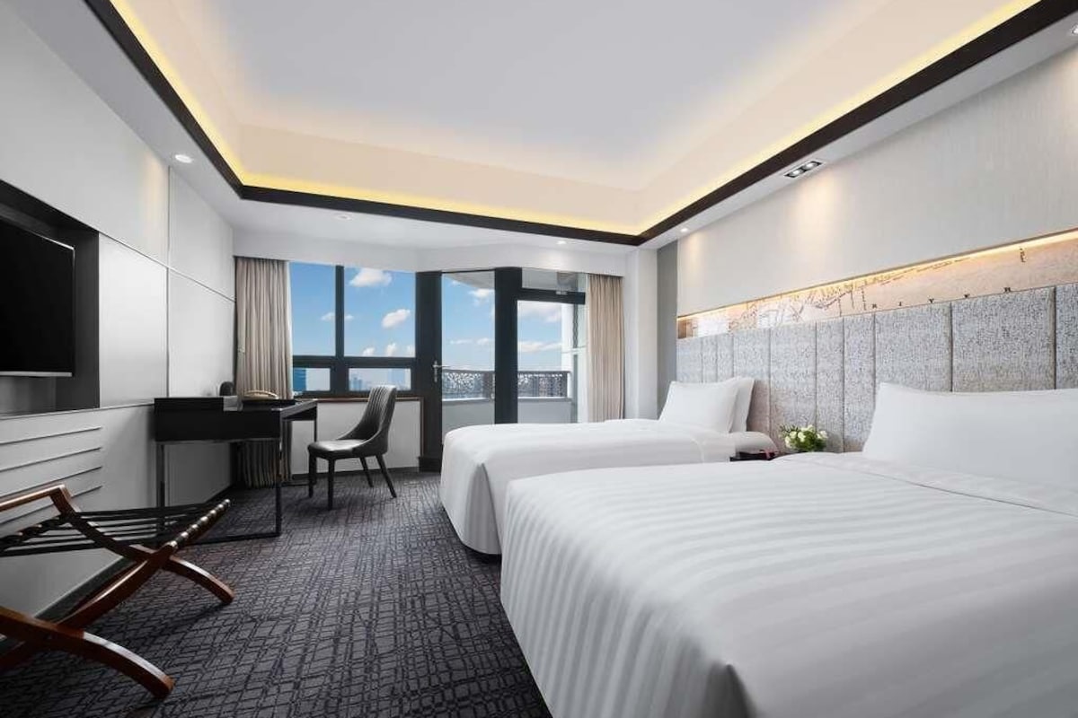 Ein Hotelzimmer mit zwei Betten, einem Schreibtisch, einem Sessel, einem Fernseher und einem großen Fenster.