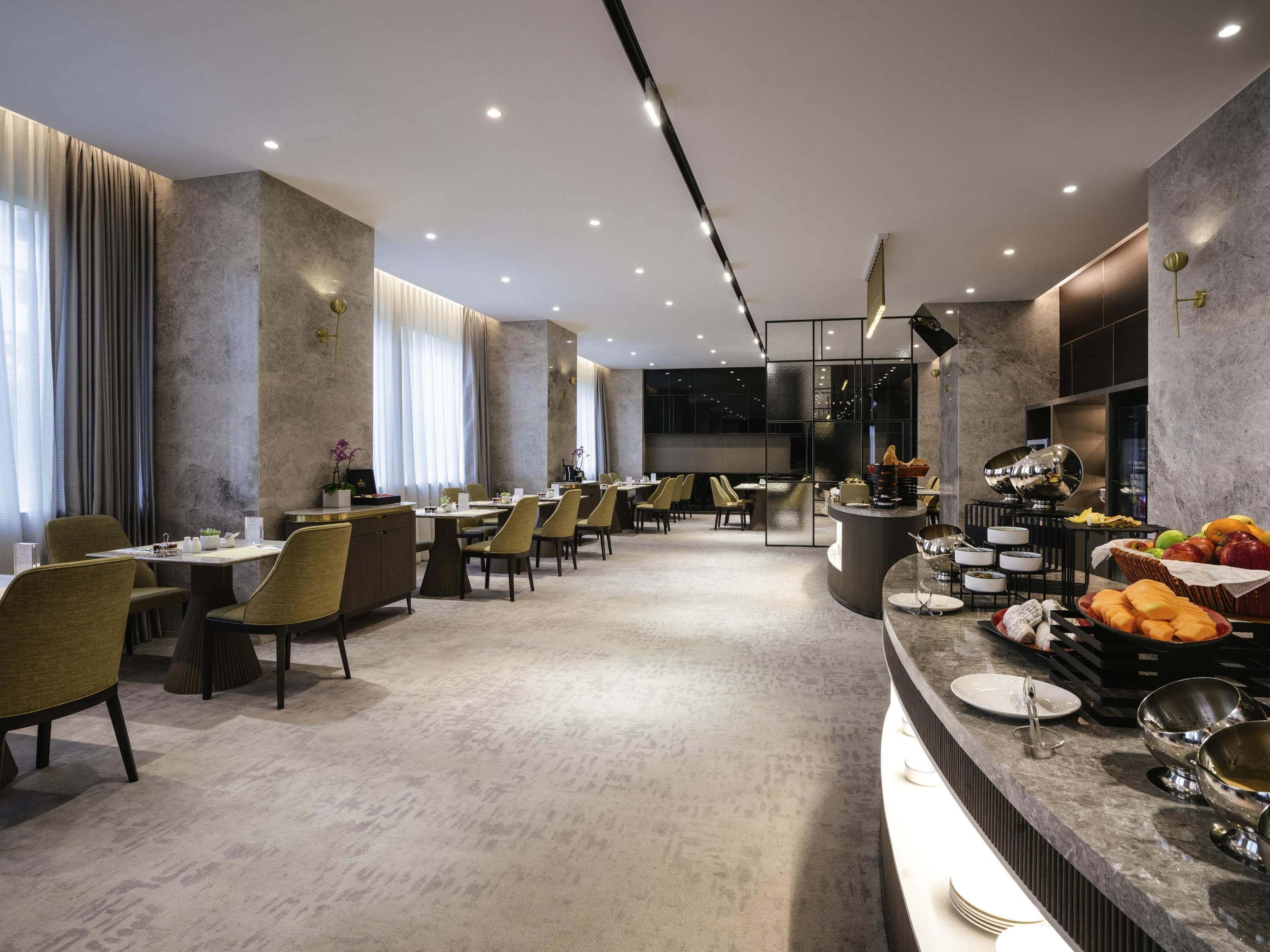 A modern hotel dining area with multiple tables and chairs, a central bar area with a fruit display, and a large window providing natural light.