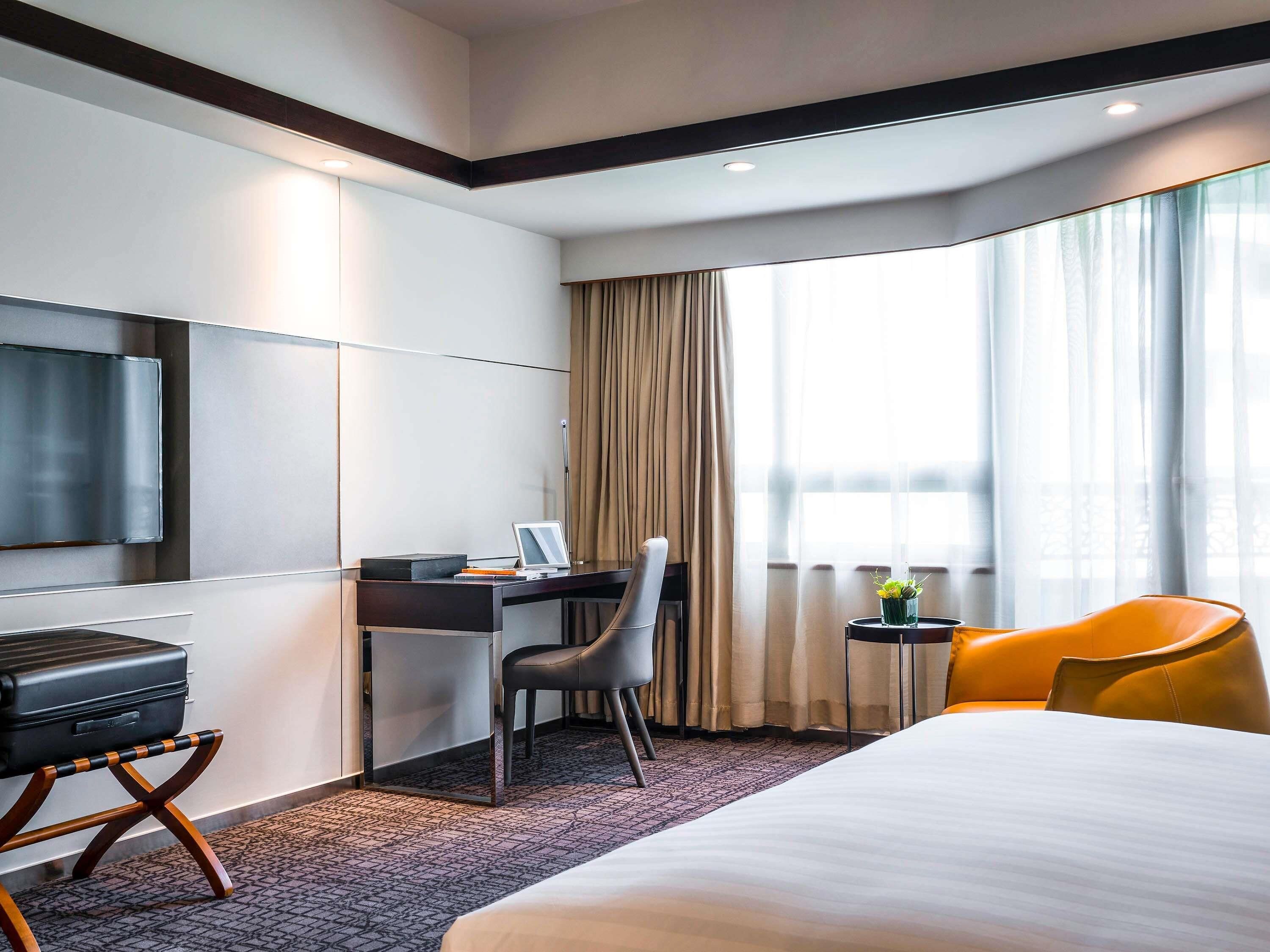 Une chambre d’hôtel comprenant un lit, un bureau avec une chaise, une télévision, une petite table avec une plante et une fenêtre avec des rideaux.