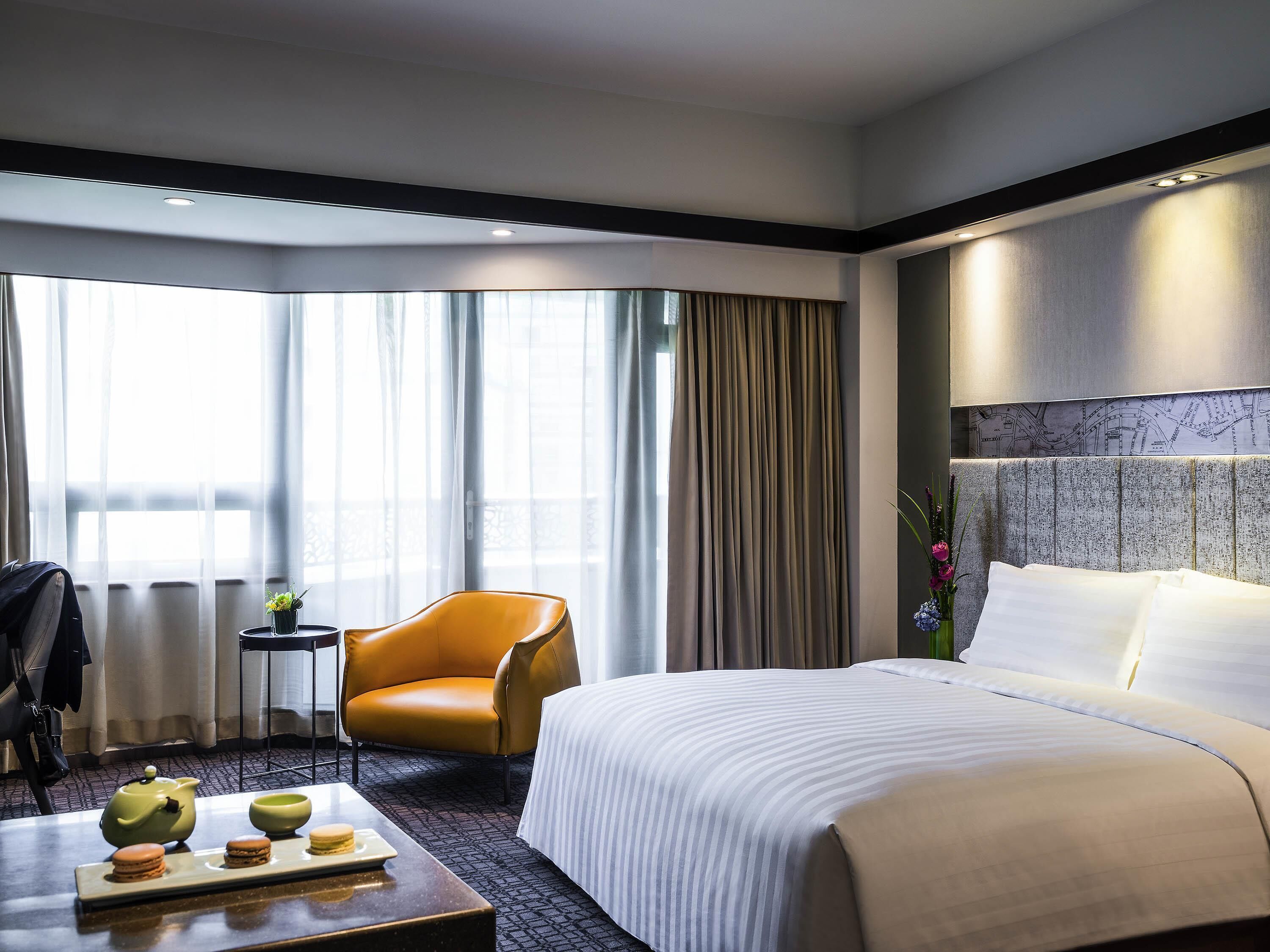 Une chambre d’hôtel avec un lit, une chaise, une petite table sur laquelle se trouvent une théière et des pâtisseries, et une vue sur l’extérieur grâce à de grandes fenêtres.