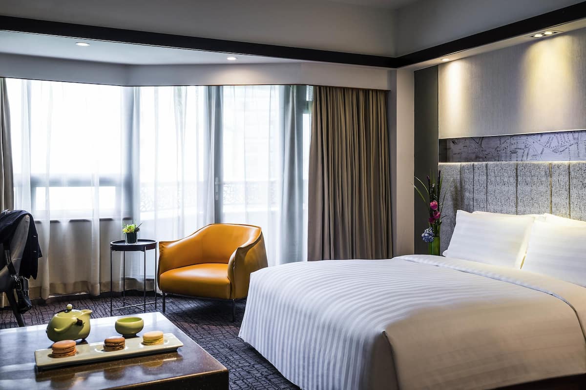 Une chambre d’hôtel avec un lit, une chaise, une petite table sur laquelle se trouvent une théière et des pâtisseries, et une vue sur l’extérieur grâce à de grandes fenêtres.