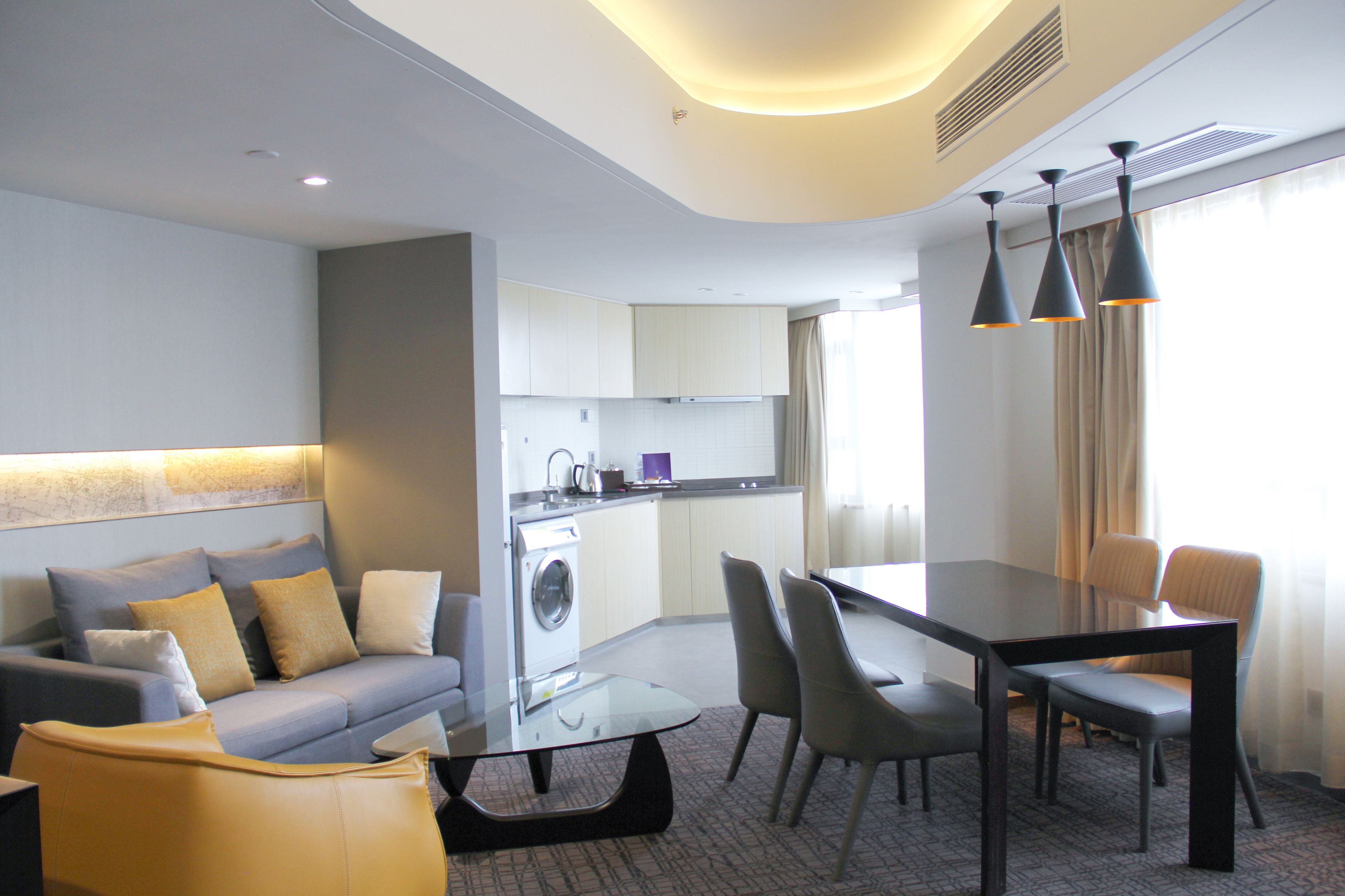 Una habitación de hotel moderna con sofá, una mesa de comedor de vidrio, sillas, lavadora y zona de cocina.