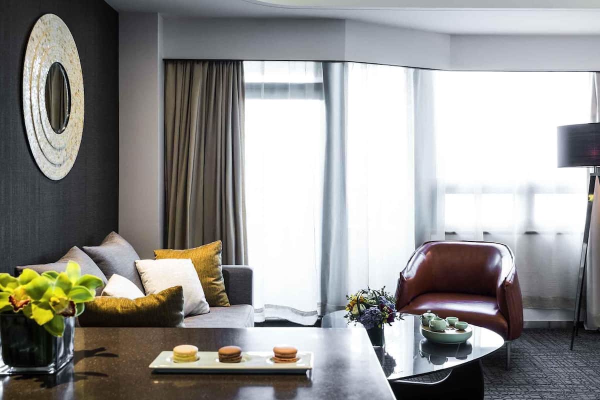 Een moderne hotelkamer met een bank, een leren fauteuil, een bijzettafeltje met gebak, een vaas met bloemen en een groot raam met gordijnen.