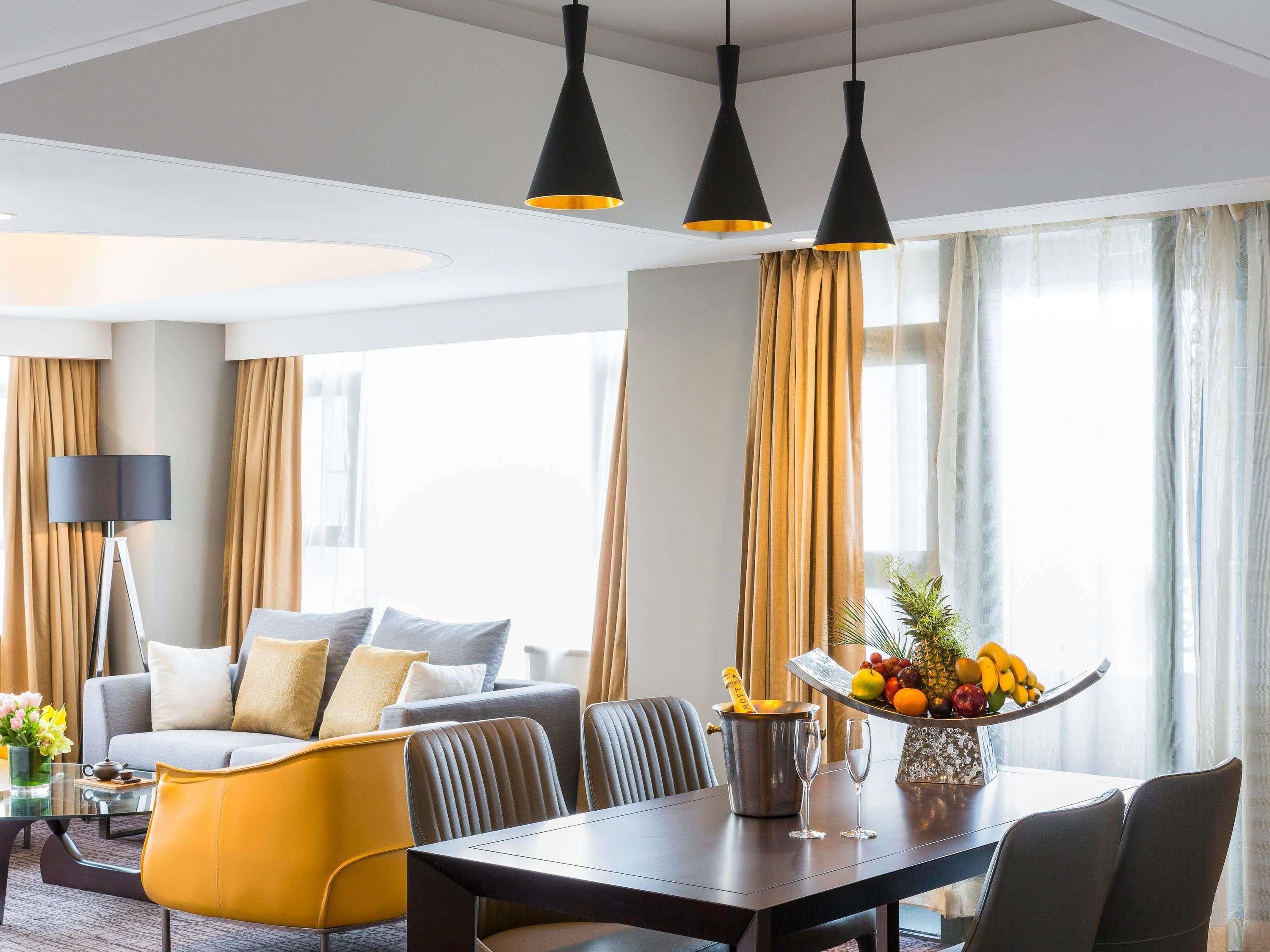 A hotel room with a dining area, a sofa, a lamp, and a fruit bowl on the table.