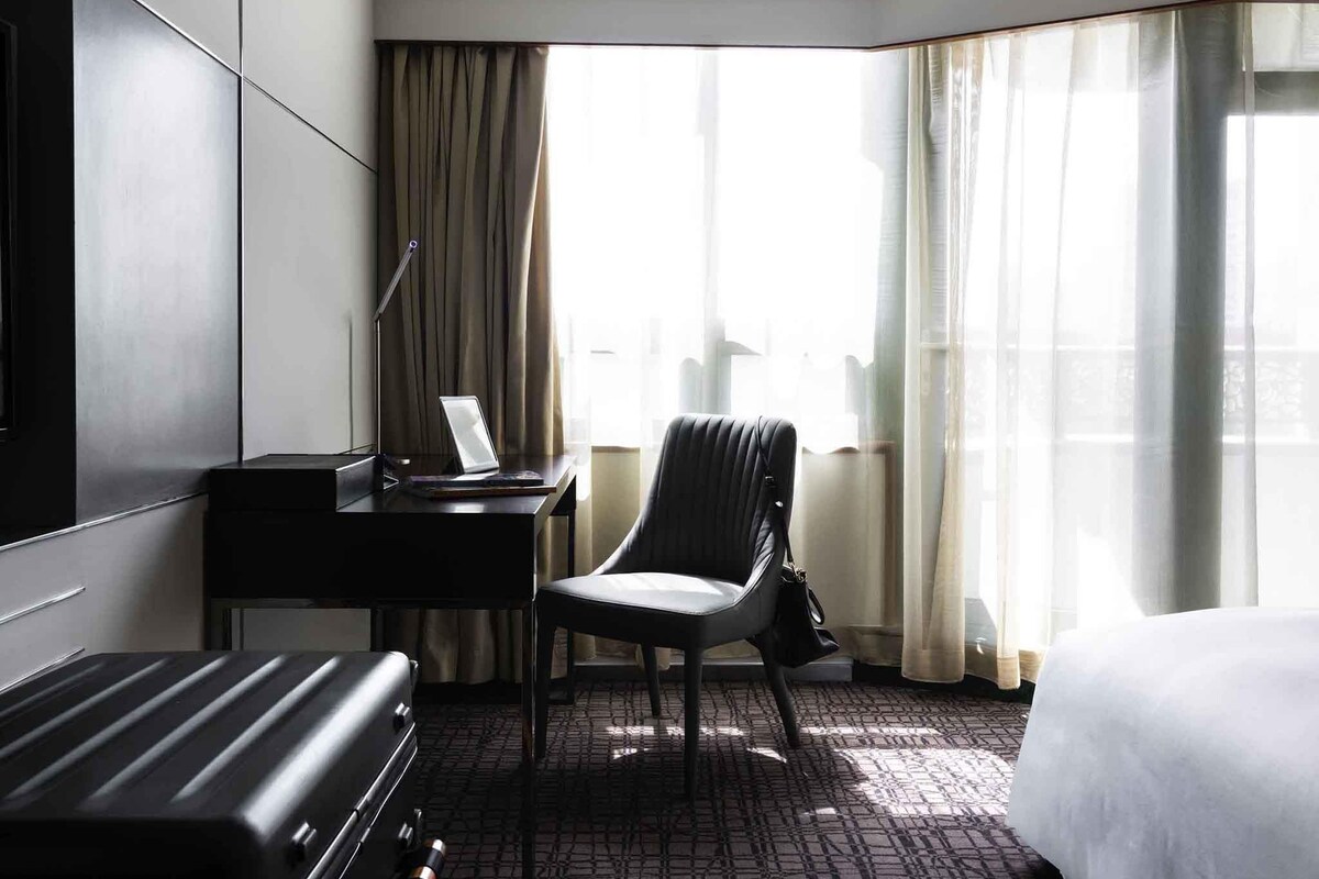A hotel room with a bed, a desk with a laptop, a chair, and a suitcase.