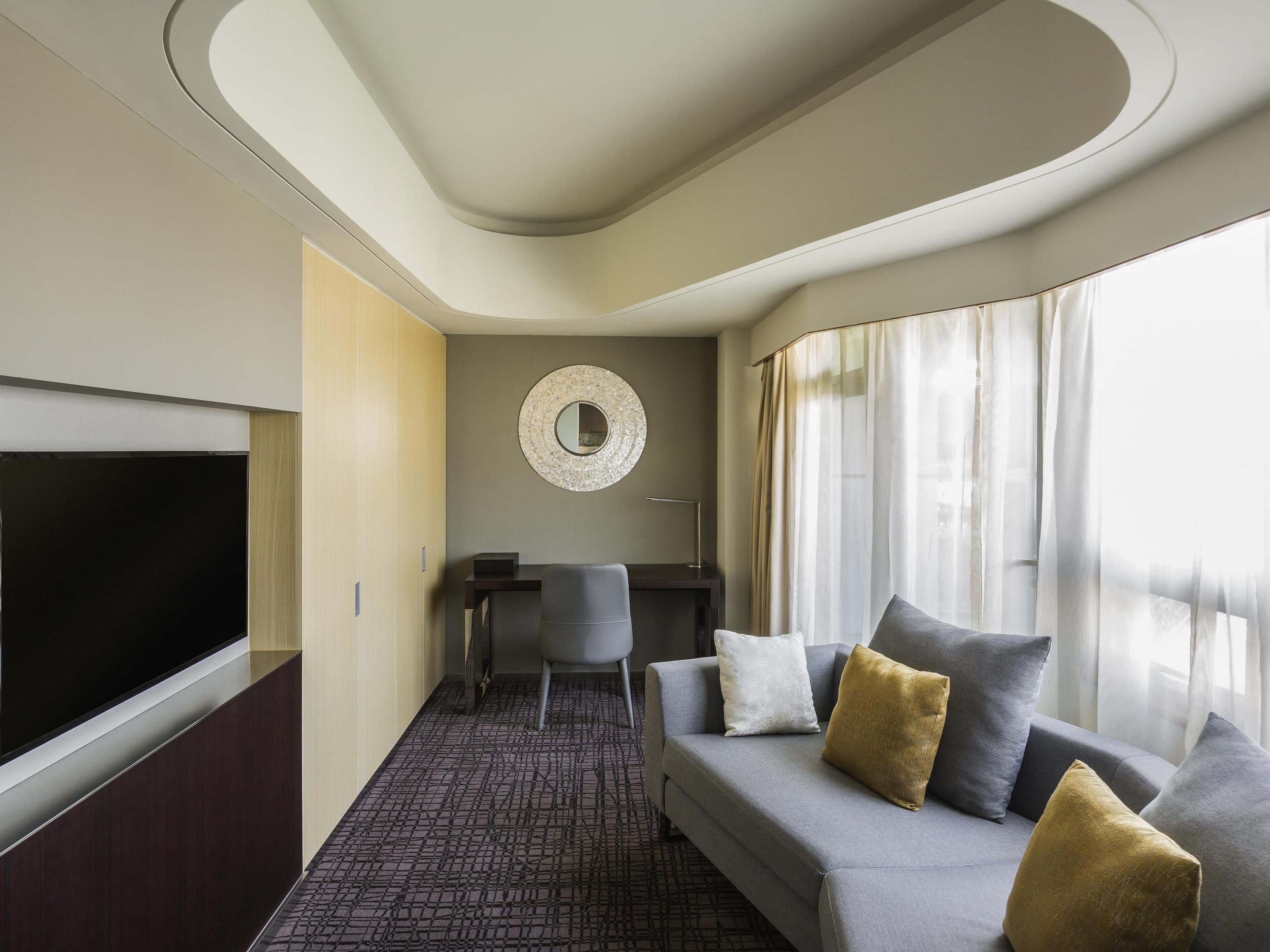 Une chambre d’hôtel moderne équipée d’un canapé, d’une télévision, d’un bureau et d’un miroir rond fixé au mur.