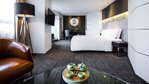 A modern hotel room with a bed, a desk, a chair, a sofa, and a coffee table with cups.