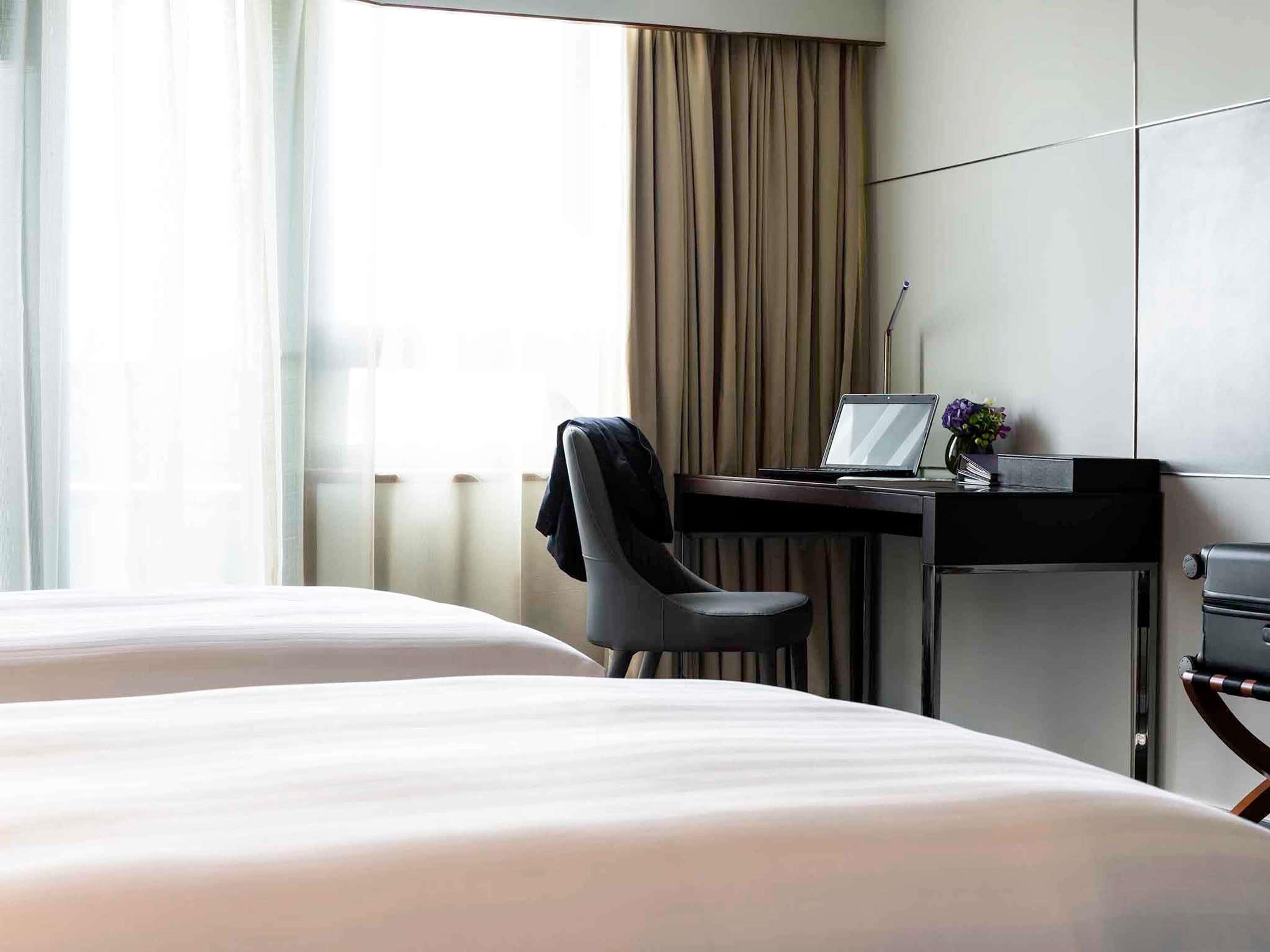 Habitación de hotel con dos camas, un escritorio con una laptop, una silla y vistas al exterior a través de ventanas amplias.