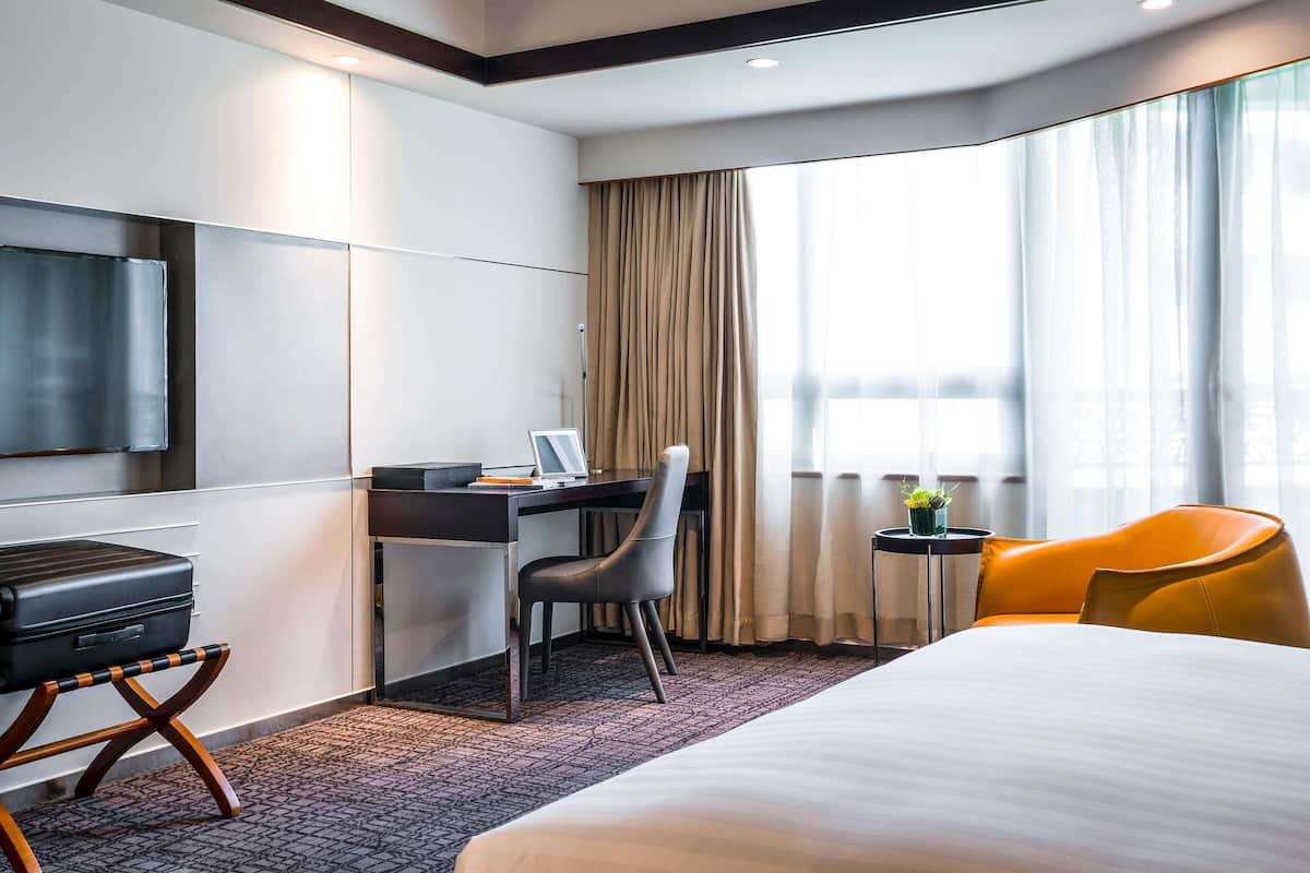 Une chambre d’hôtel comprenant un lit, un bureau avec une chaise, une télévision, une petite table avec une plante et une fenêtre avec des rideaux.