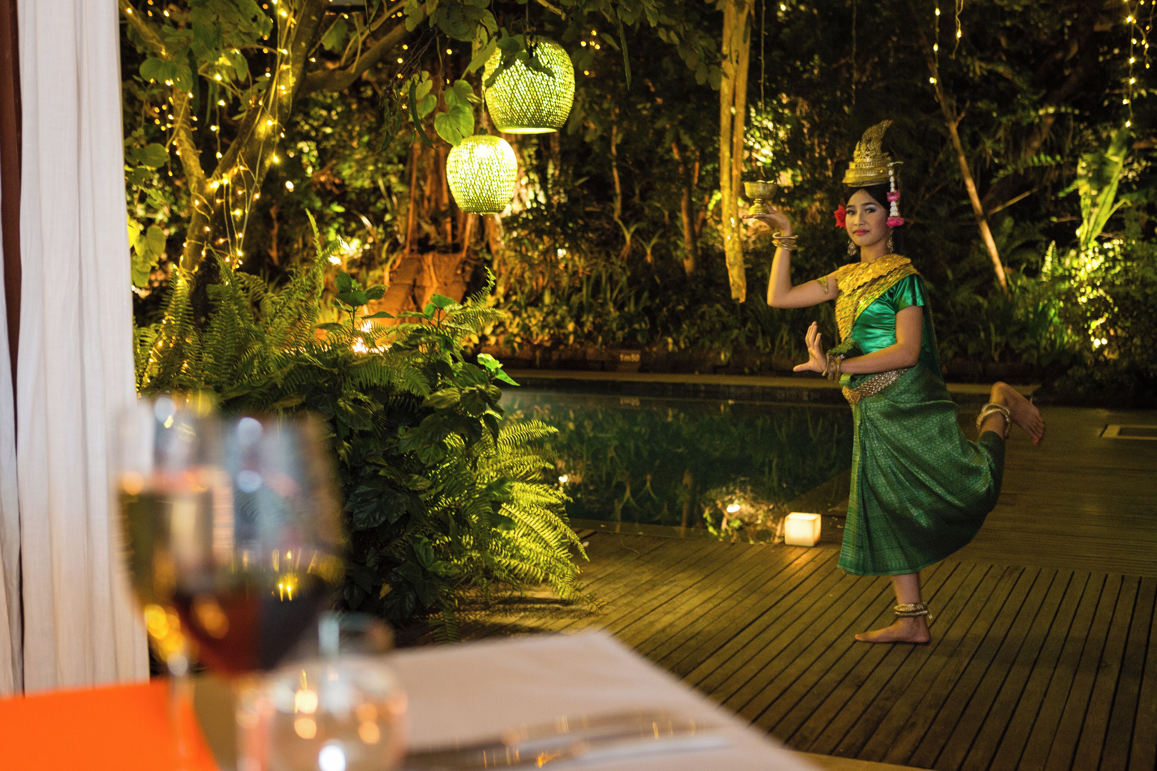 A person in traditional attire standing on a wooden deck with greenery and string lights in the background.