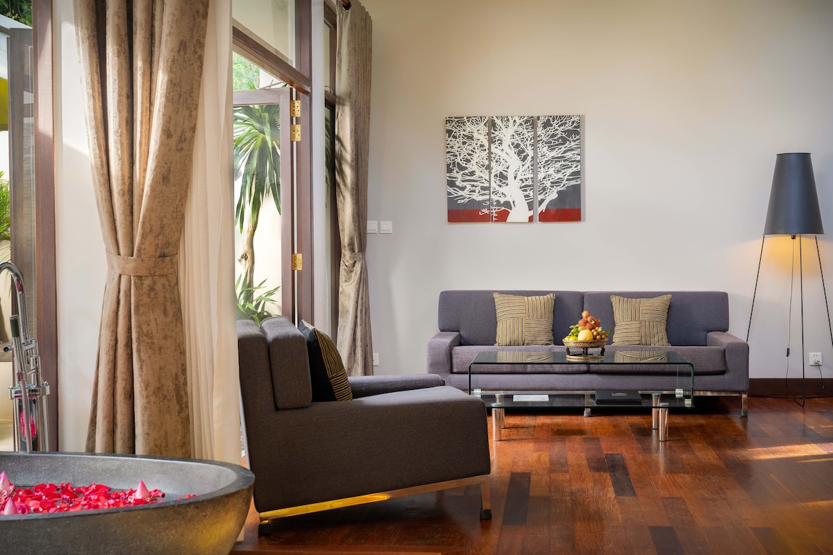 Een moderne woonkamer met een grijze bank, een glazen salontafel en een schilderij van een boom aan de muur.