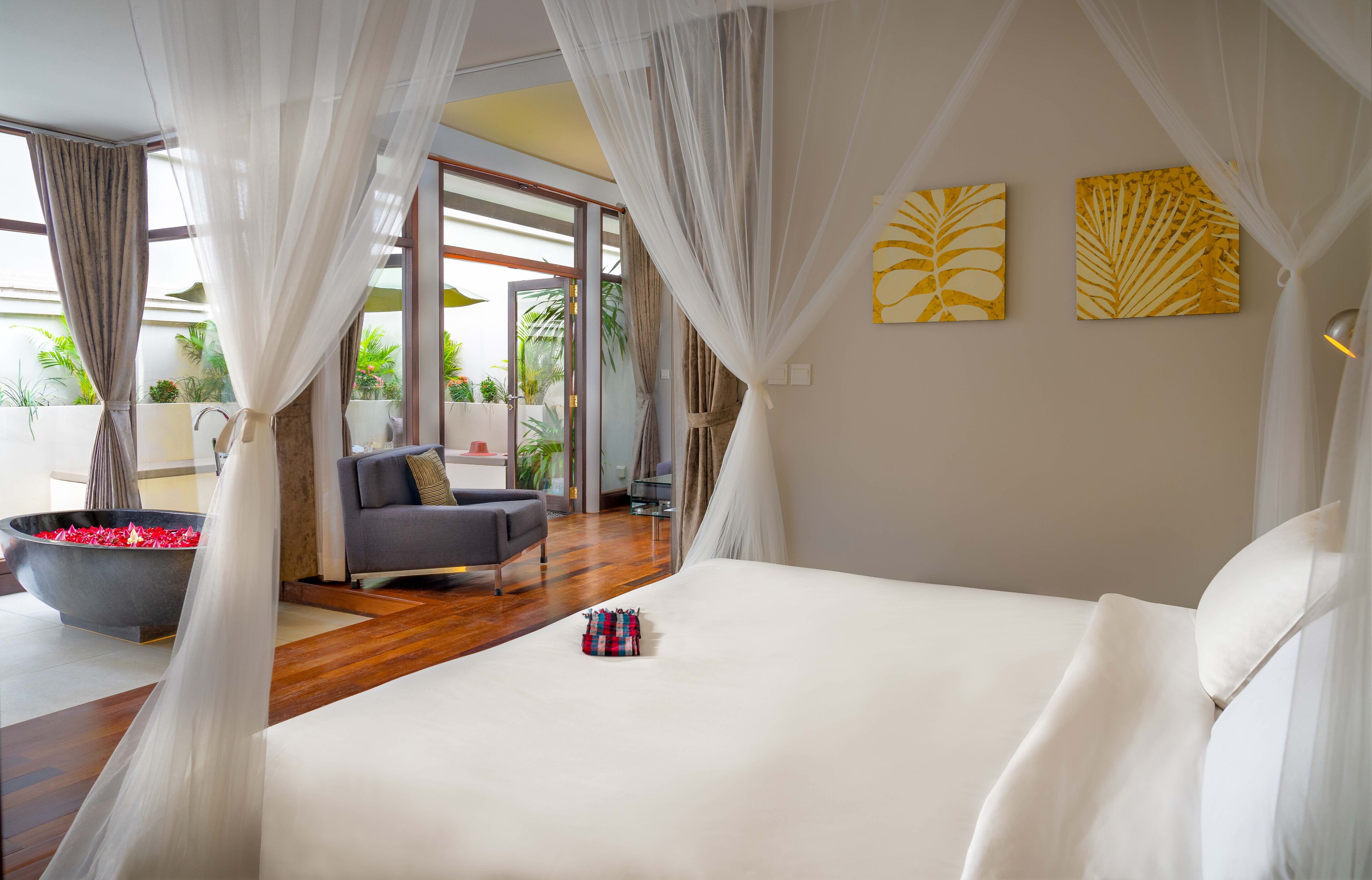 Un dormitorio con una cama grande, un sillón gris, una mesita pequeña y un cuenco grande lleno de flores rojas.