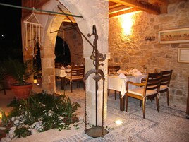Un área de comedor al aire libre con paredes de piedra, mesas y sillas de madera. Hay plantas en macetas y una lámpara colgante.
