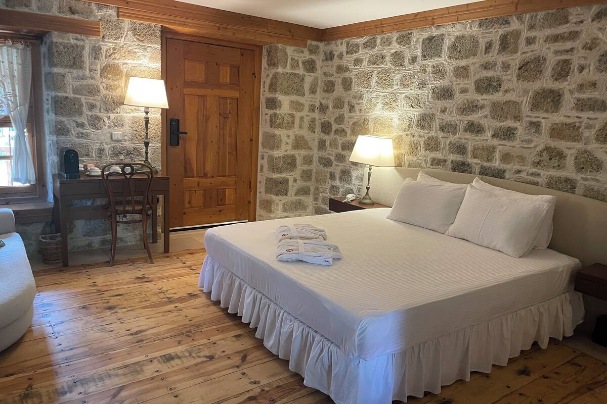 Une chambre avec un mur en pierre, un plancher en bois, un lit avec une literie blanche, un bureau avec une lampe et une porte en bois.