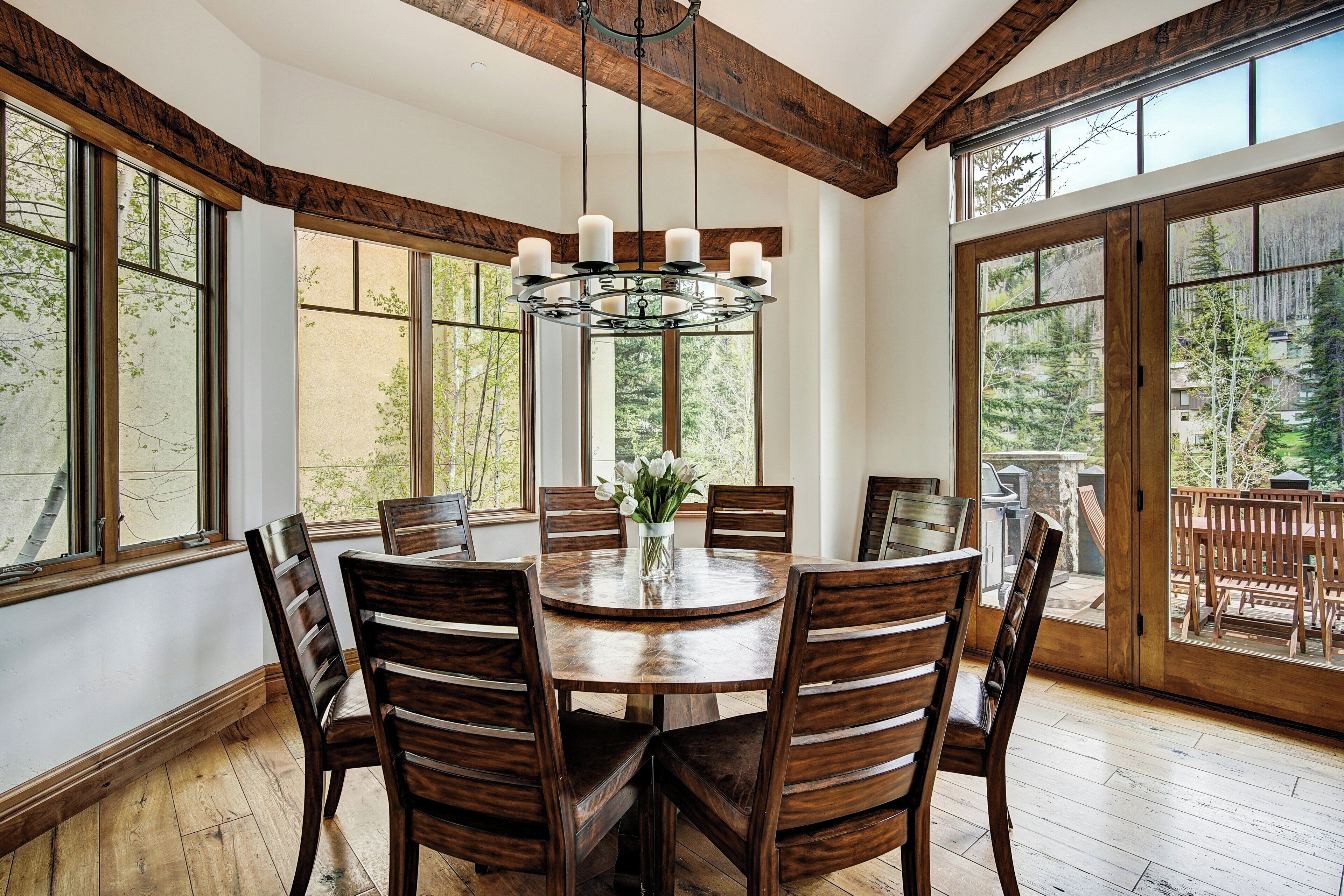 A dining room with a wooden table and chairs, large windows, and a chandelier.