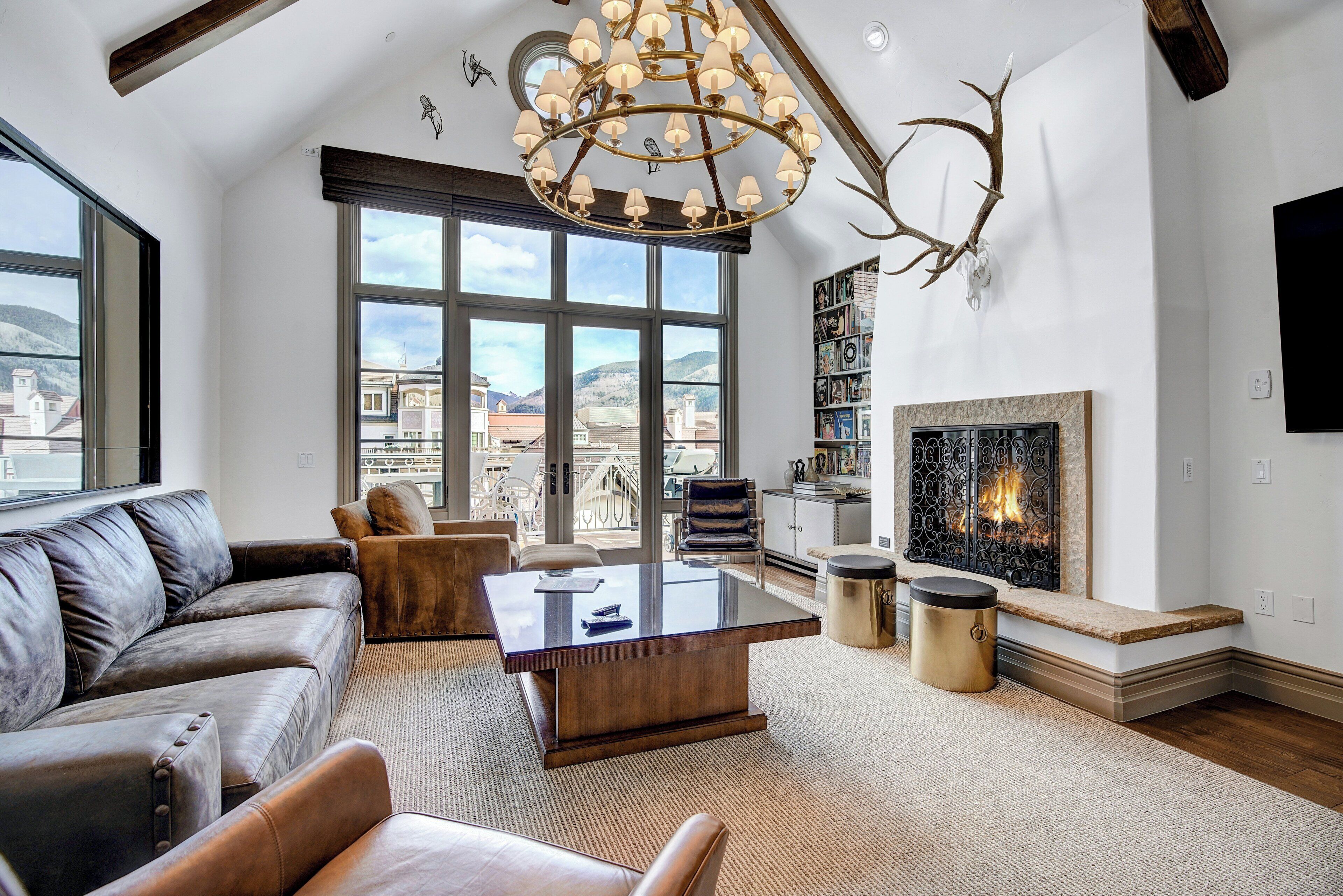 A spacious living room with a large window, a fireplace, a leather sofa, and a modern chandelier.