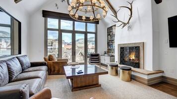 A spacious living room with a large window, a fireplace, a leather sofa, and a modern chandelier.