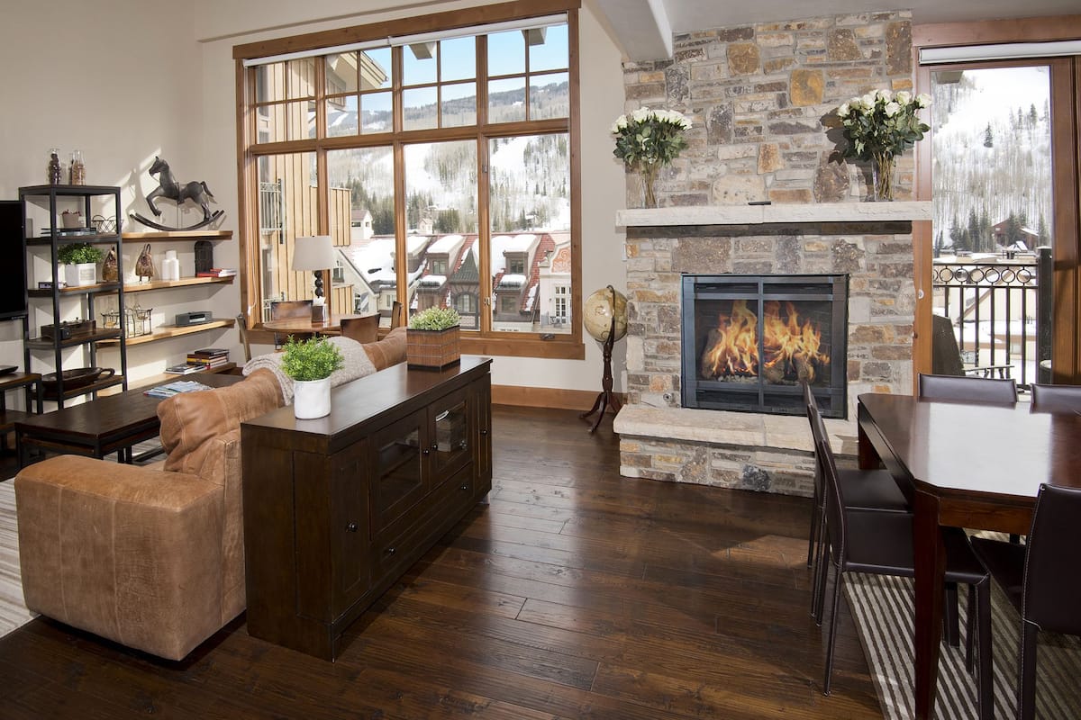 A spacious living room with a stone fireplace, wooden flooring, and large windows offering a view of a snowy landscape.