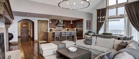 A spacious living room with a large sofa, a coffee table, and a chandelier. There is a kitchen area with wooden cabinets and a refrigerator. The room has a high ceiling with exposed beams and large windows with curtains.