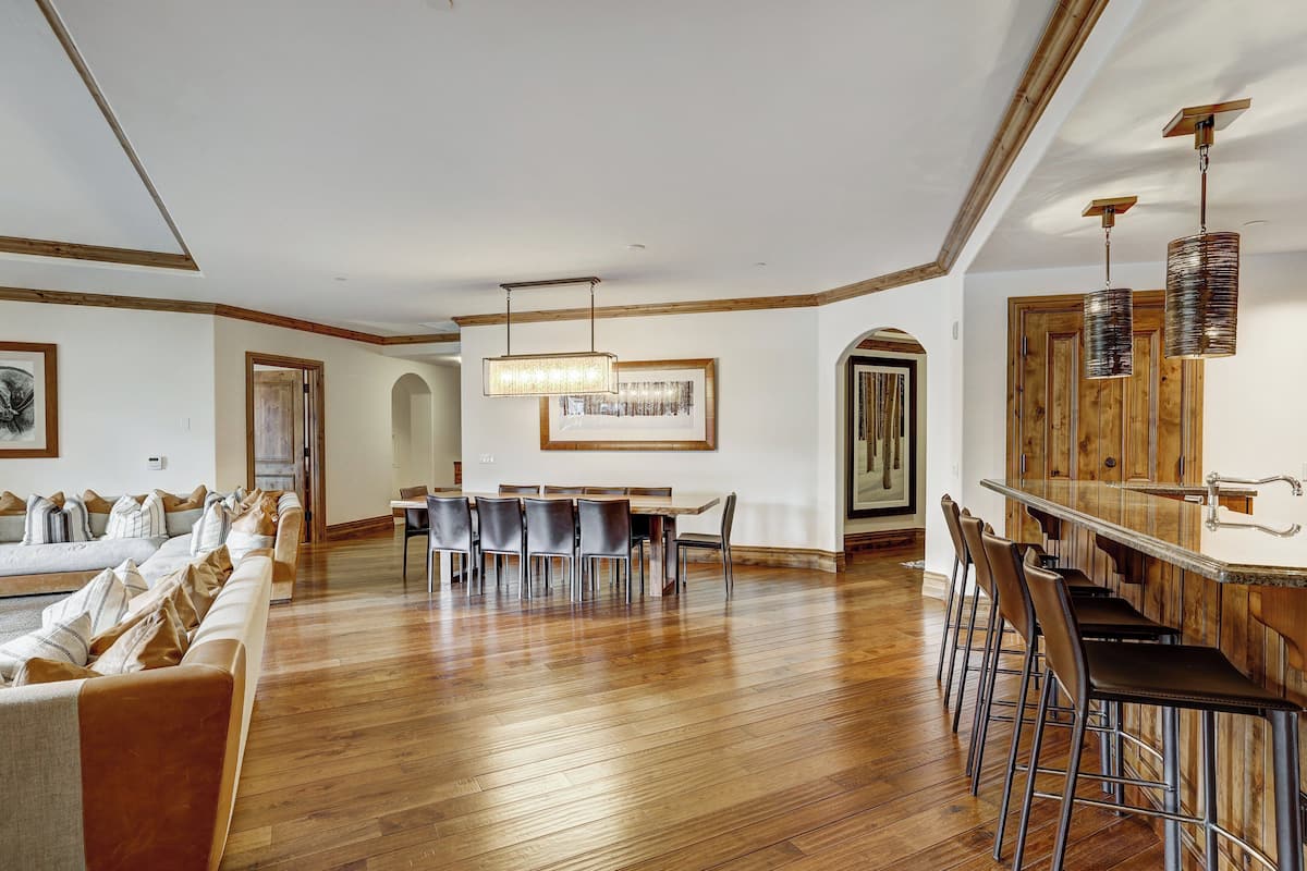 A spacious living area with a wooden floor, a dining table with chairs, a bar area with stools, and a large window providing natural light.