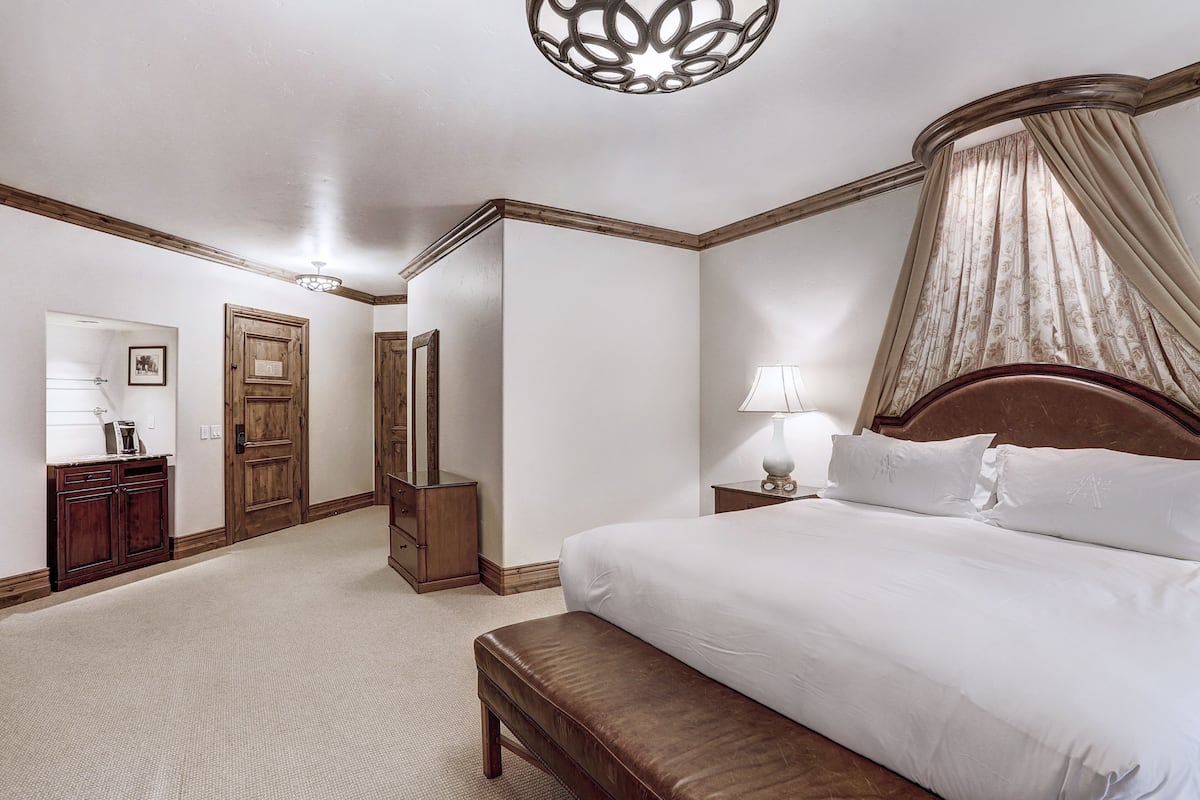A hotel room with a large bed, a bedside table with a lamp, a wooden bench, a wardrobe, and a mirror.