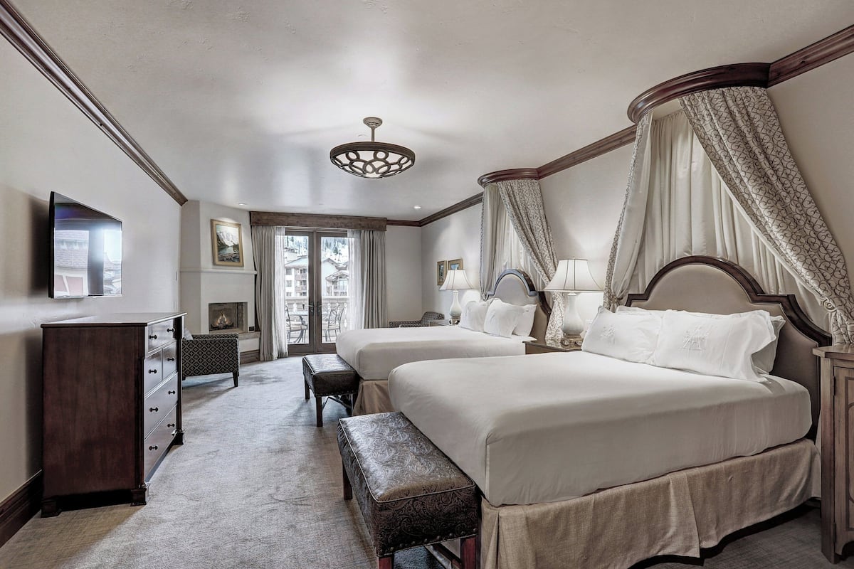 A hotel room with two beds, a desk, a chair, a bench, and a television.